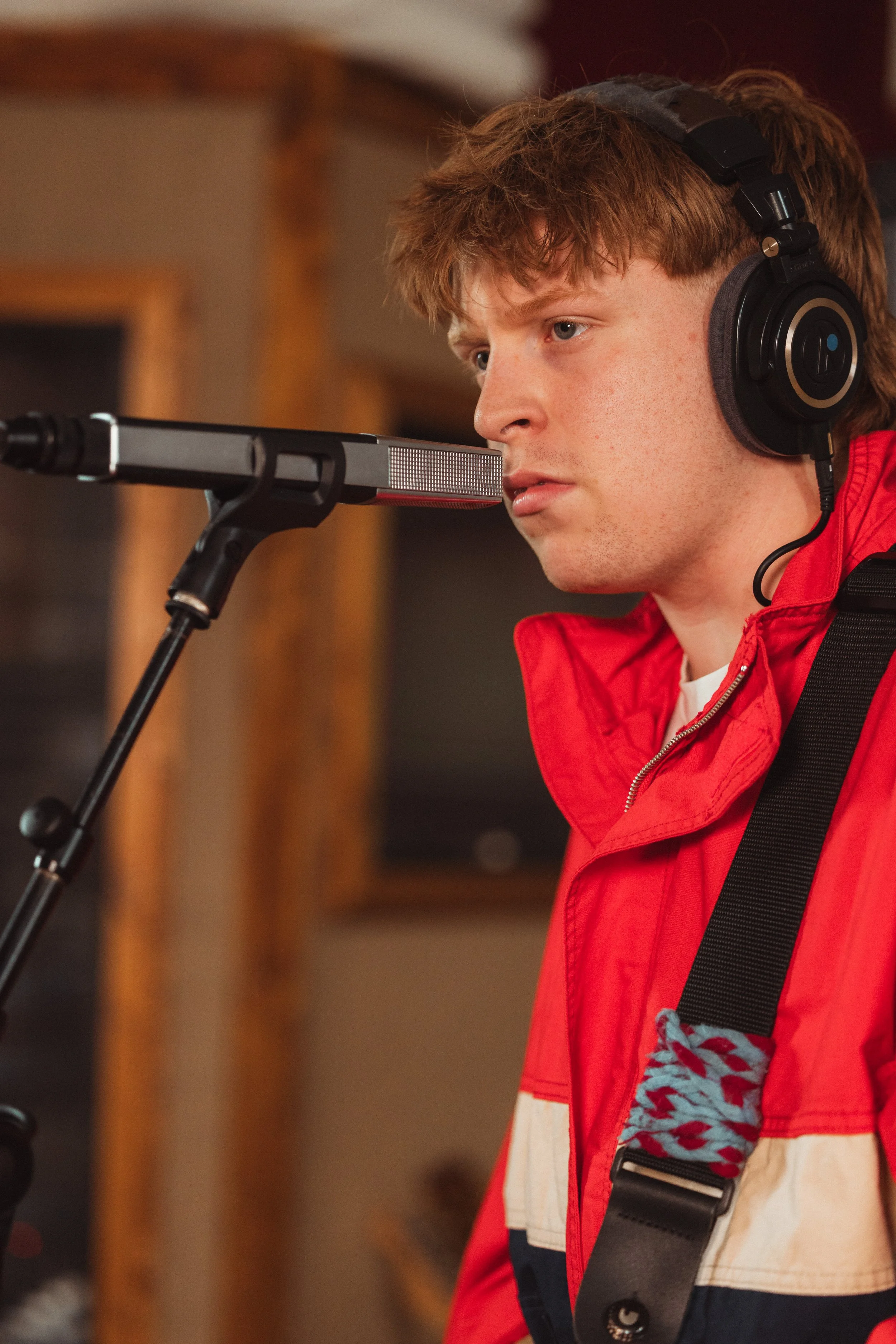 A young man with reddish-brown hair and blue eyes, wearing black over-ear headphones and a red, white, and blue jacket, is focused and singing or speaking into a microphone in a recording studio.