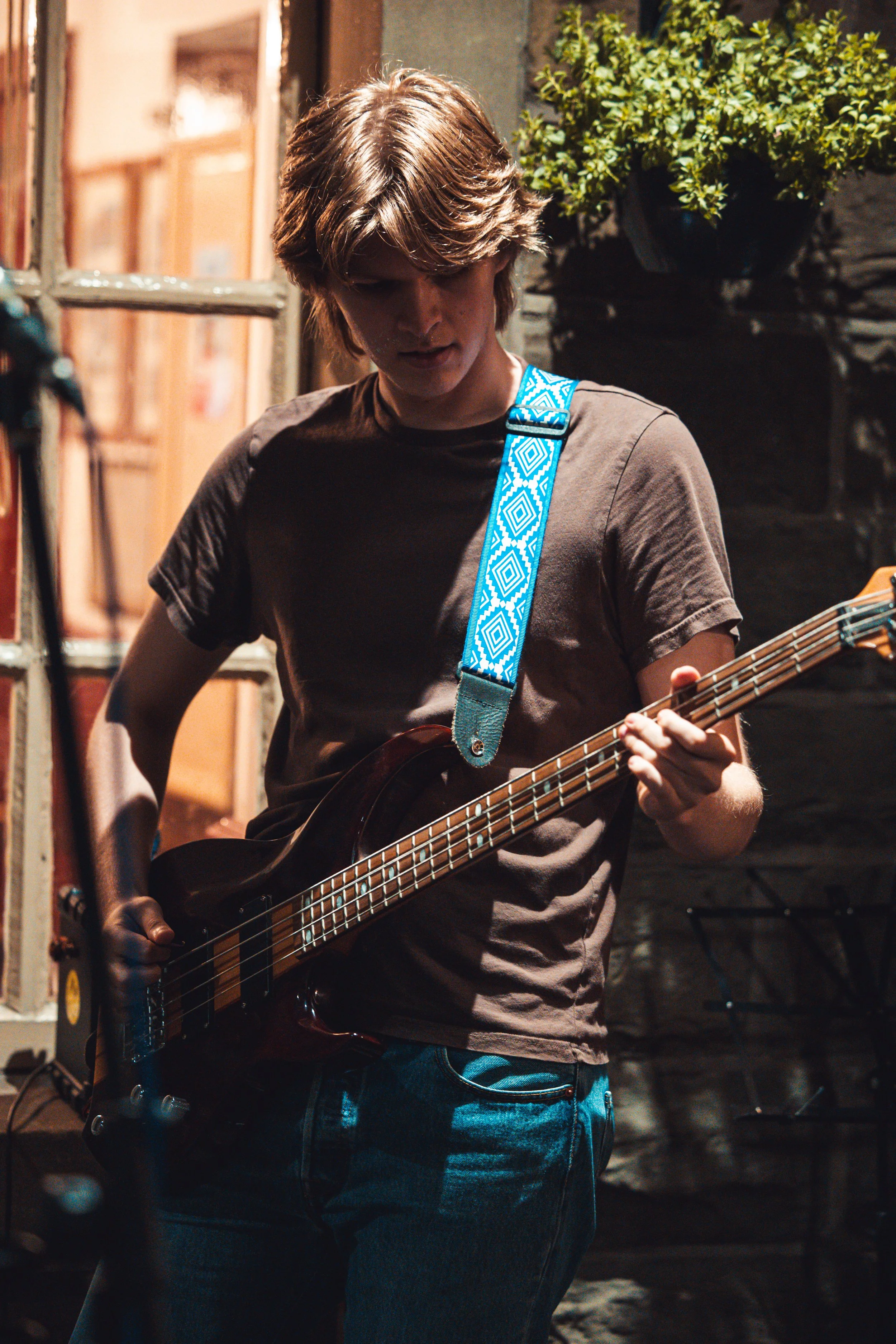 A young man with medium-length blonde hair plays an electric guitar in a dimly lit indoor setting. He's wearing a brown t-shirt and blue jeans, with a blue patterned guitar strap. The background includes a brick wall, window, and a potted plant.