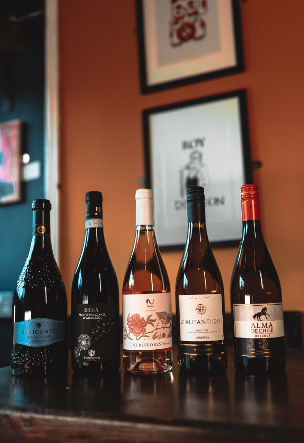 Five bottles of wine on a wooden table, with artwork hanging on a reddish wall in the background.