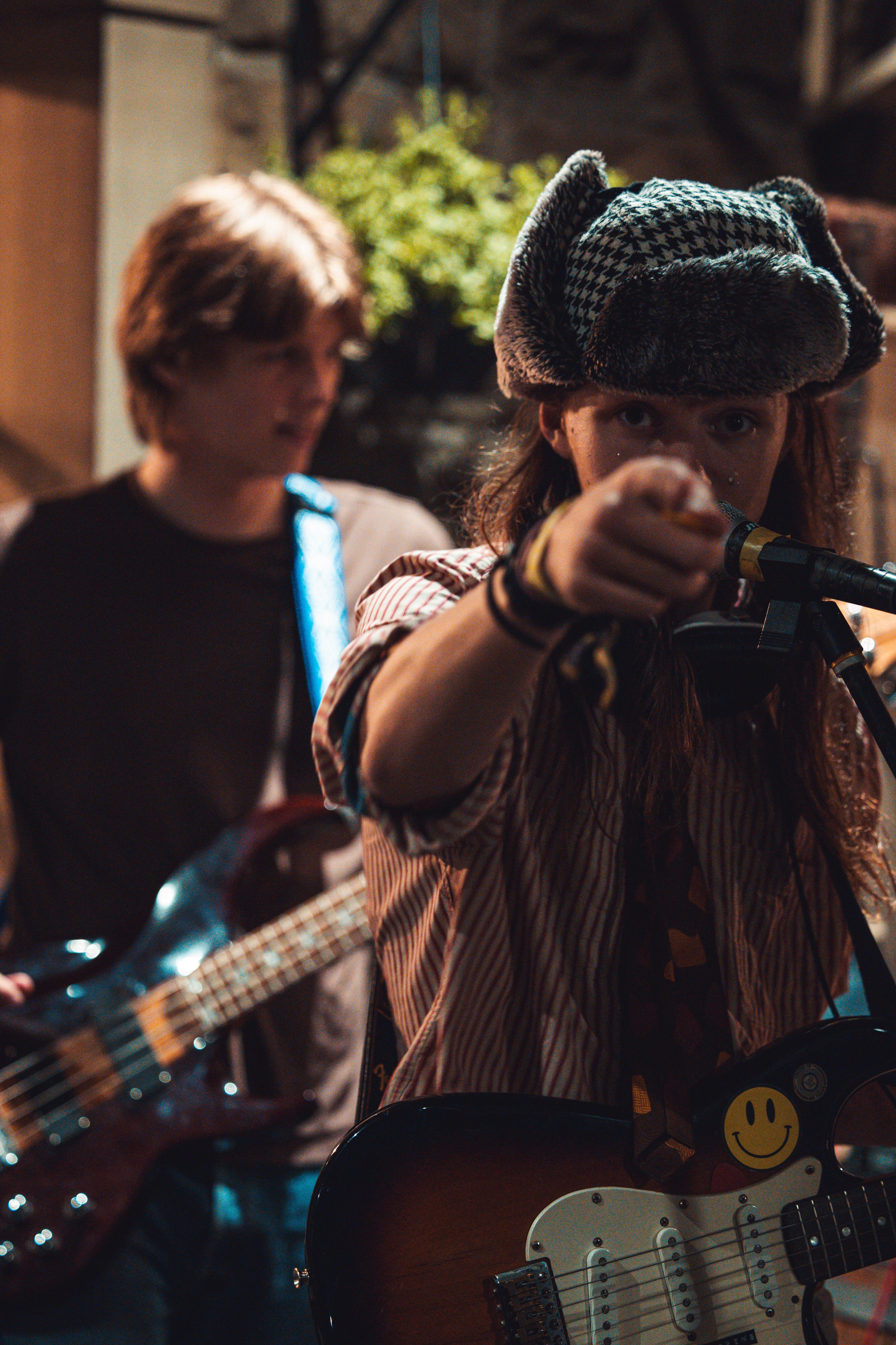 Young girl with a houndstooth hat playing guitar and pointing at the camera, with another person blurred in the background.