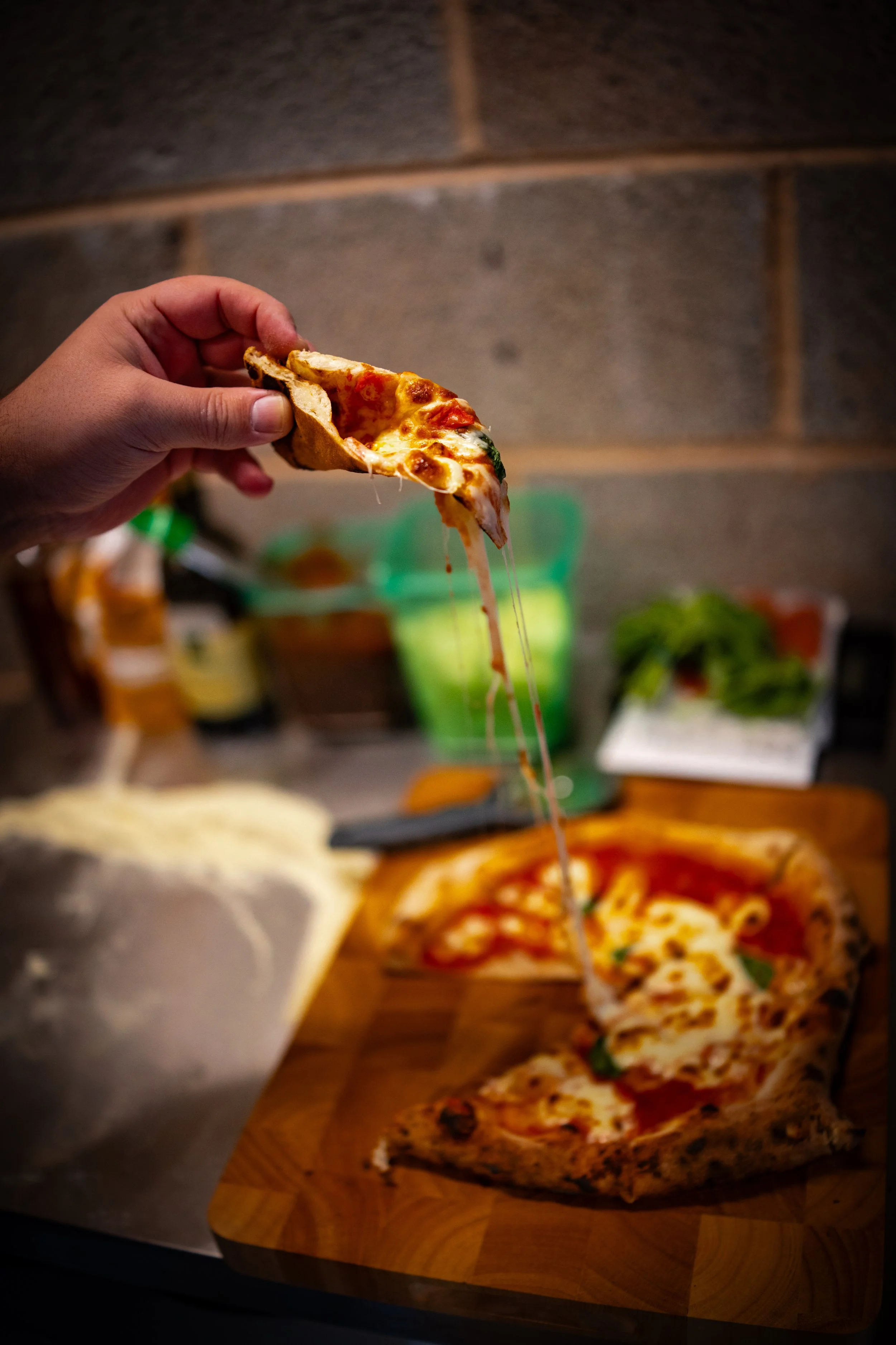 A hand holding a slice of pepperoni pizza with melted cheese, with a pizza box on a wooden board and a cluttered kitchen counter in the background.