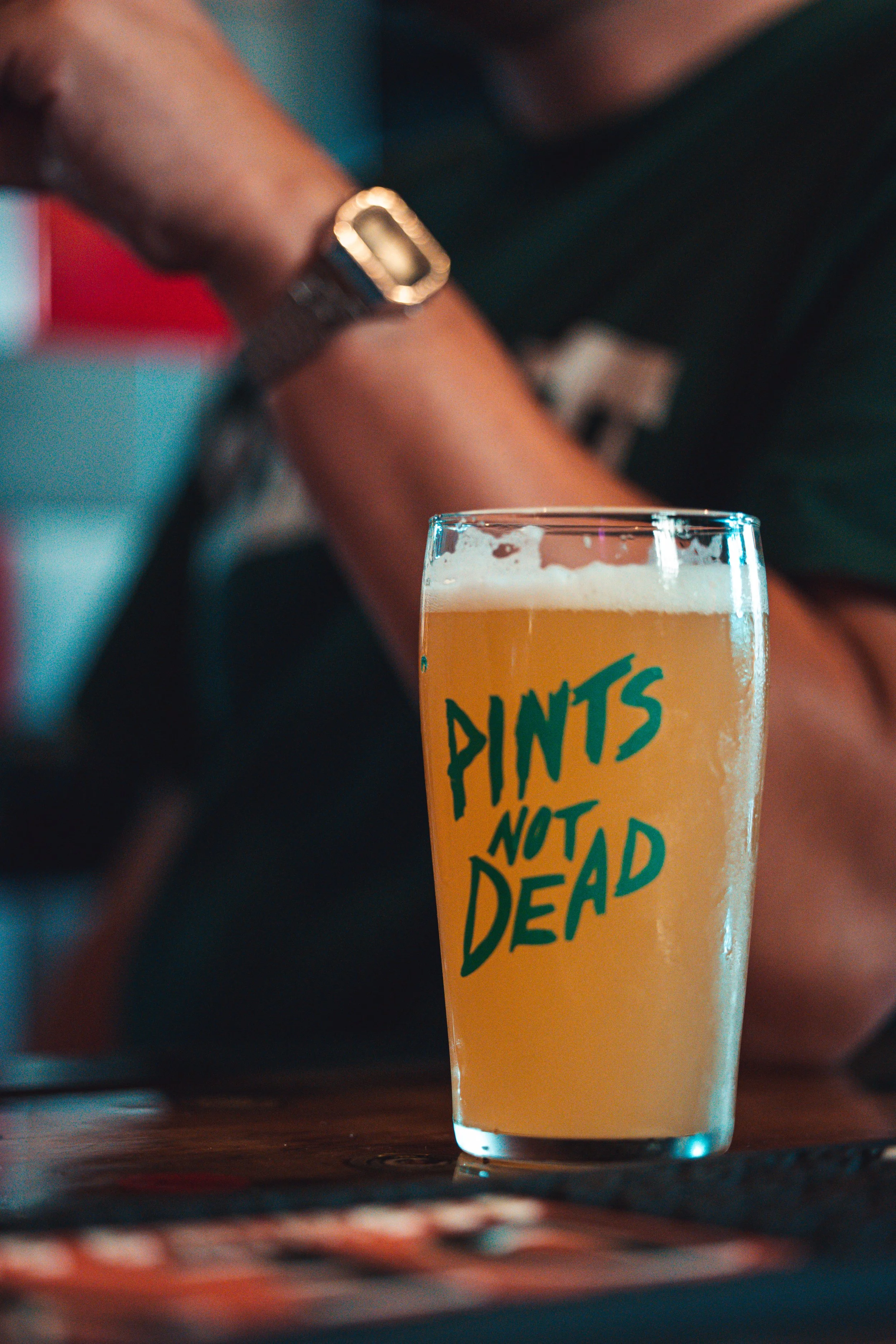 A close-up of a glass of beer on a table, with the glass featuring the phrase 'PINTS NOT DEAD' written in bold green letters. In the background, a person is wearing a watch and a dark-colored shirt.