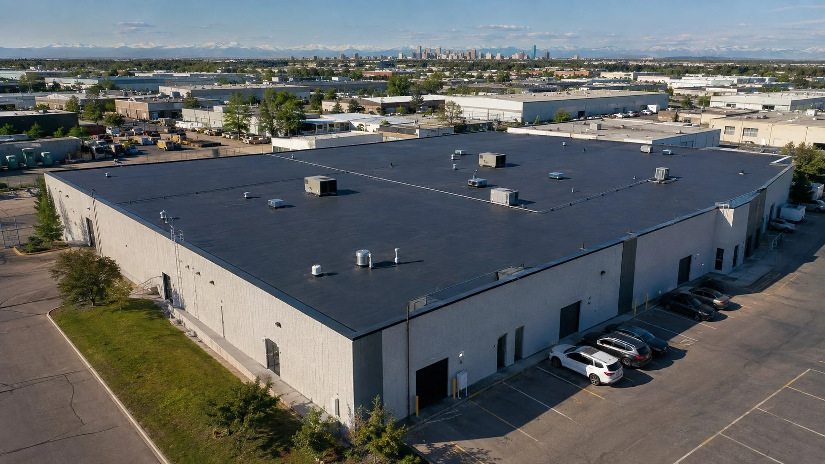 An aerial view of a large commercial building with a flat black roof, surrounded by a parking lot with several cars, and other similar warehouses in the background under a blue sky.