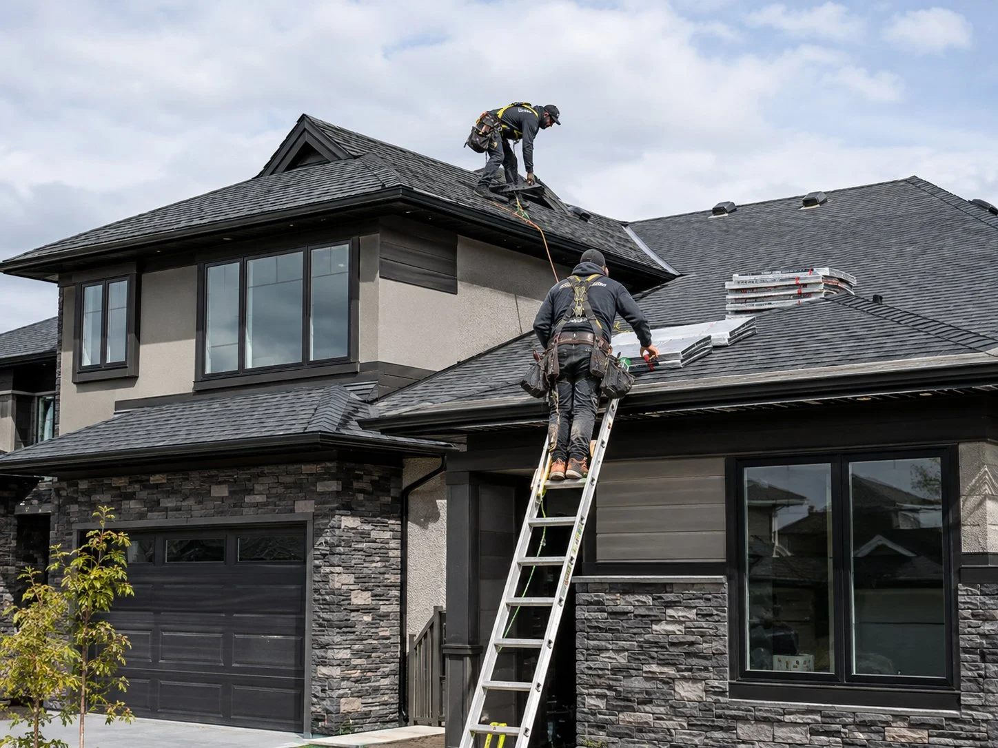 Two workers on a ladder repairing a roof of a modern house with gray siding and stone accents, one on the roof and the other on the ladder, under a cloudy sky.