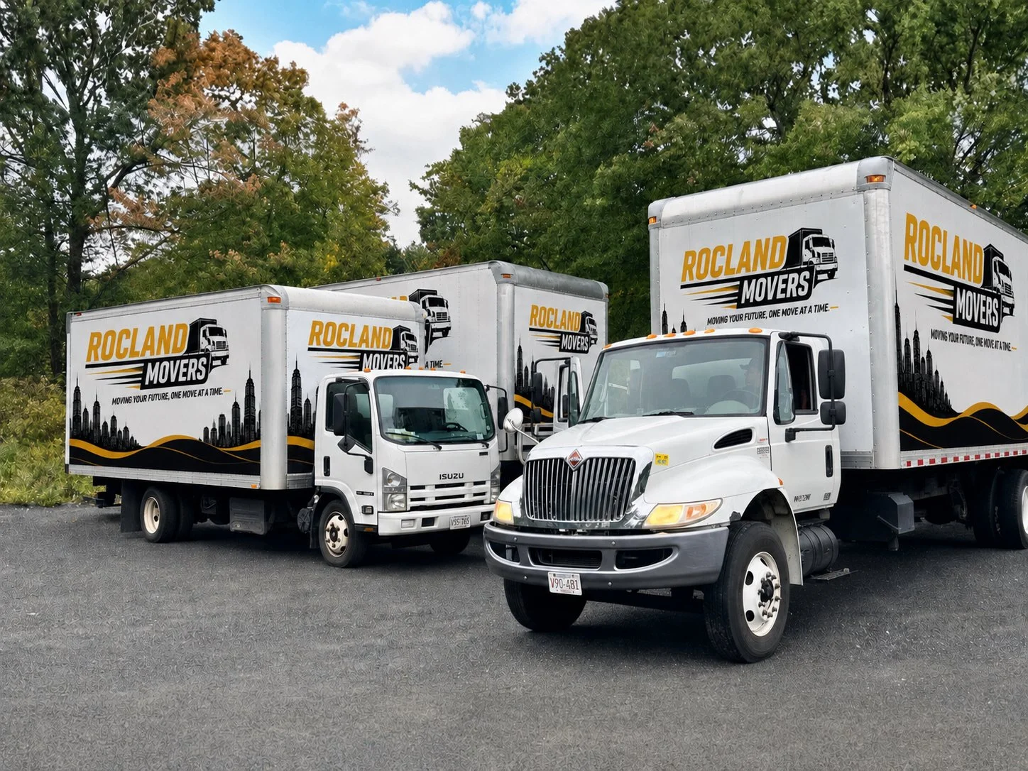 Three white moving trucks with 'ROCLAND MOVERS' logos parked side by side on a gravel pavement, surrounded by green trees and a partly cloudy sky.