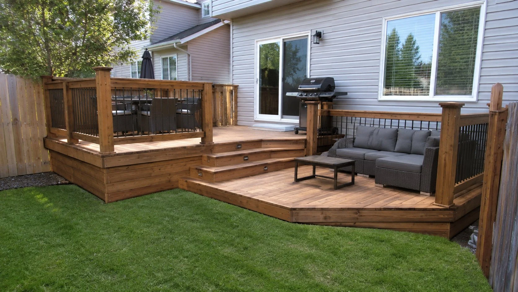 Wooden backyard deck with stairs, outdoor seating, a grill, and a sliding glass door, surrounded by a grassy yard and a wooden fence.