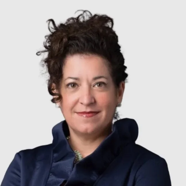 A woman with curly brown hair wearing a dark blue jacket and a necklace, smiling at the camera against a plain light background.