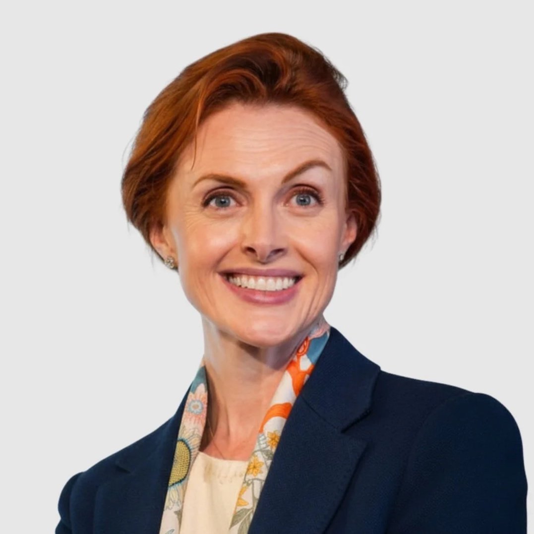 A woman with short red hair and blue eyes, smiling, wearing a navy blazer and a colorful scarf, against a light gray background.