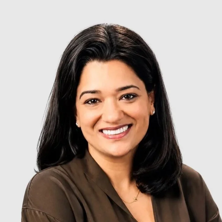 A woman with shoulder-length black hair, smiling, wearing a brown top and small earrings, against a light gray background.
