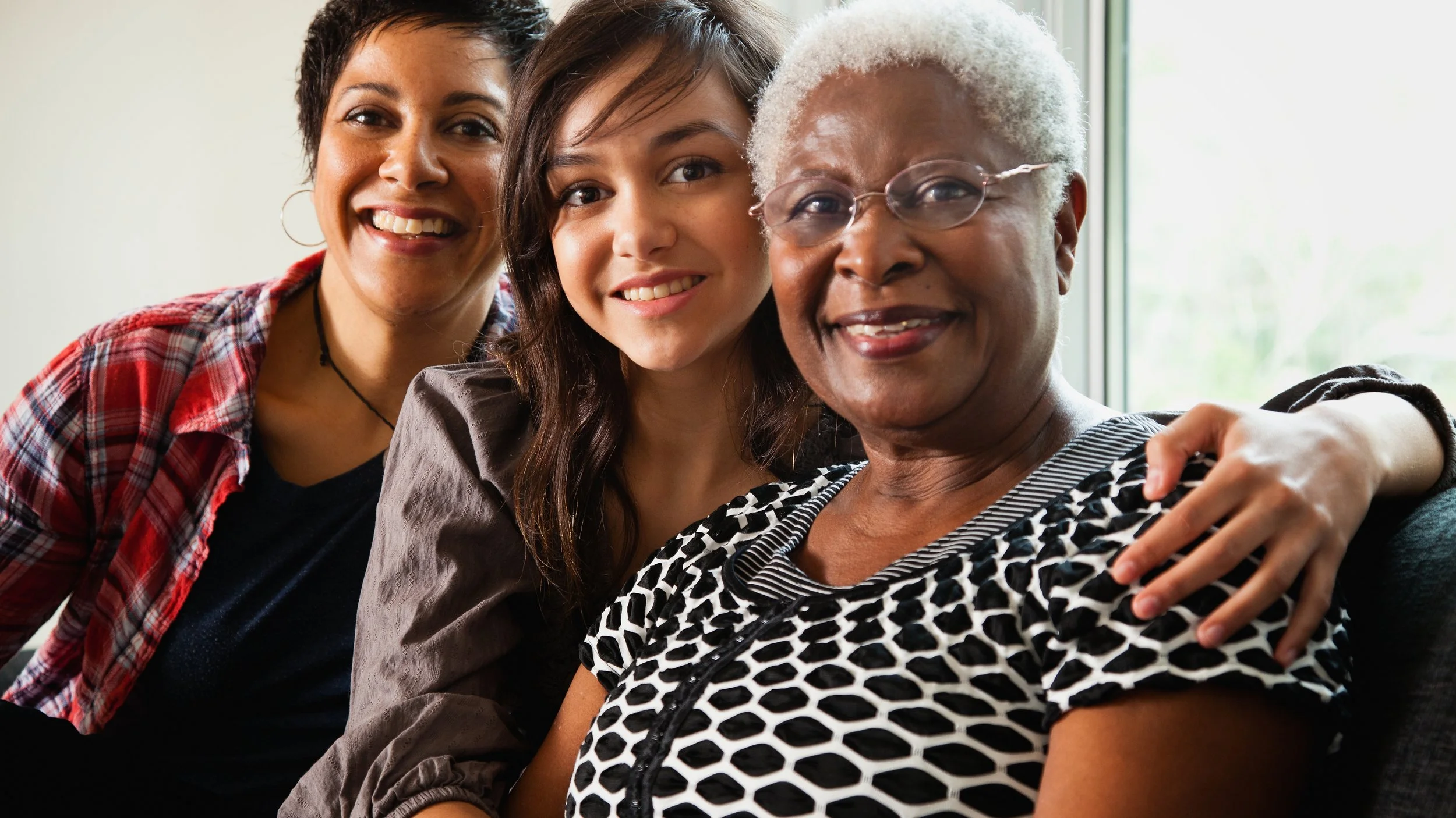 Three women of different ages smiling and sitting close together indoors near a window.