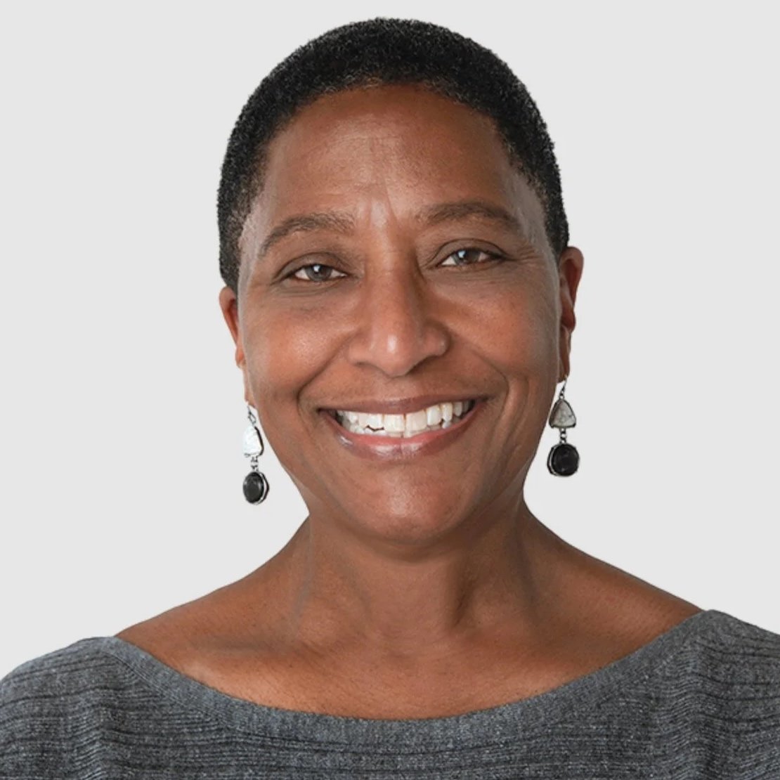 Close-up of a smiling African American woman with short hair, wearing earrings and a gray top, against a plain background.
