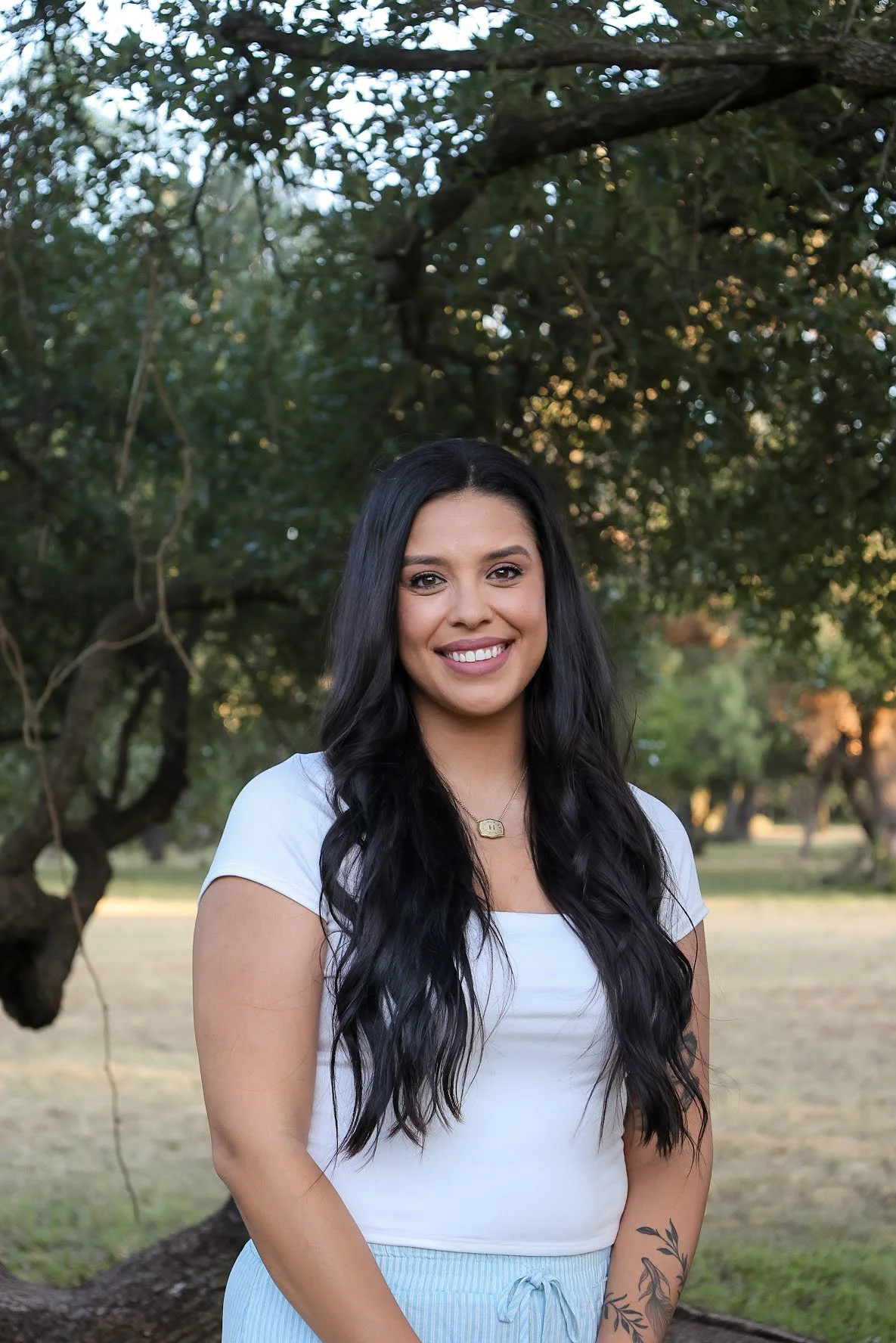A smiling woman with long black wavy hair, wearing a white t-shirt and light blue striped shorts, standing outdoors in a park with trees and greenery in the background.