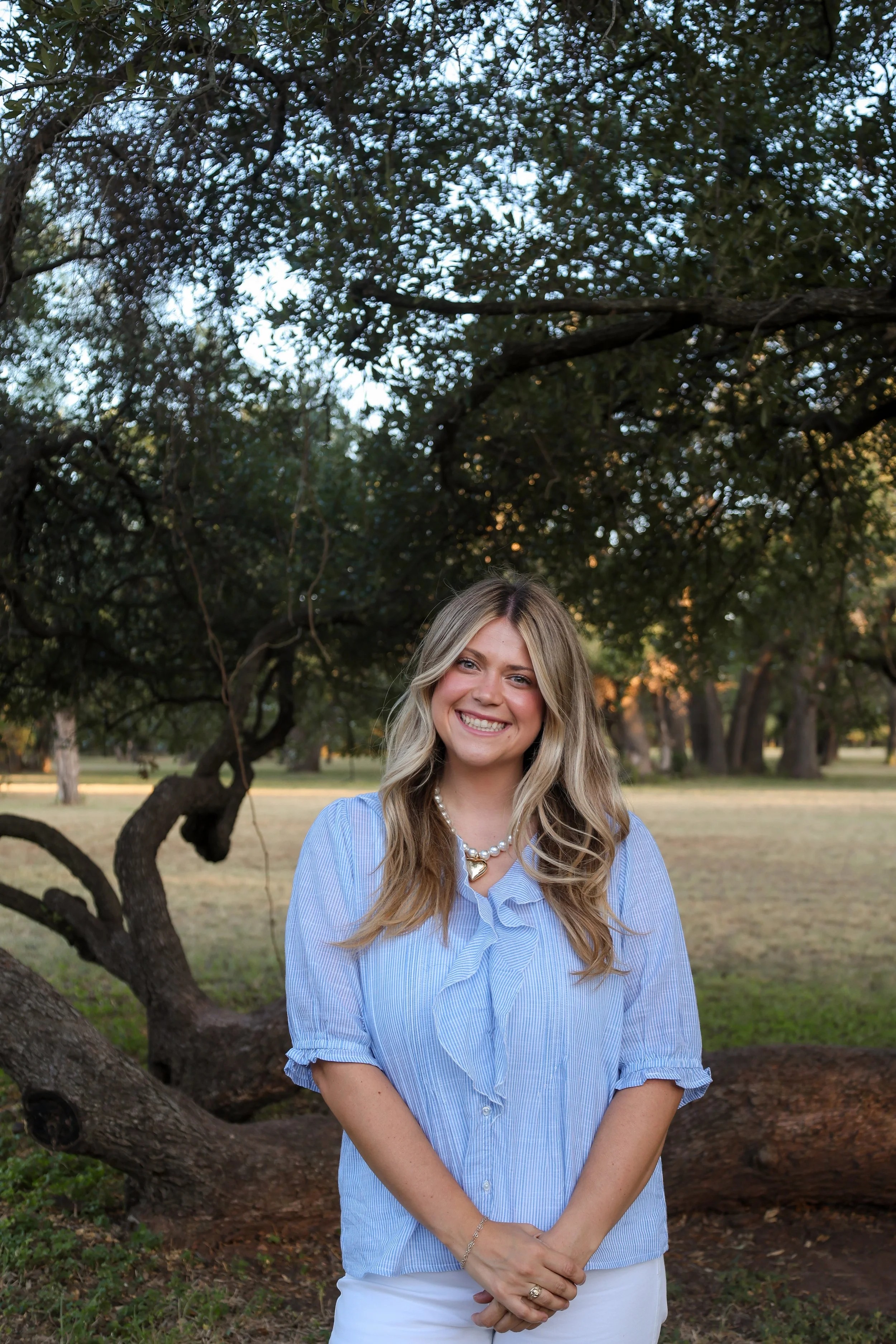 Smiling woman with blonde wavy hair, wearing a blue striped shirt with rolled-up sleeves, white pants, pearl necklace, and rings, standing outdoors in front of a tree with a sprawling branch and green foliage, during late afternoon or early evening.