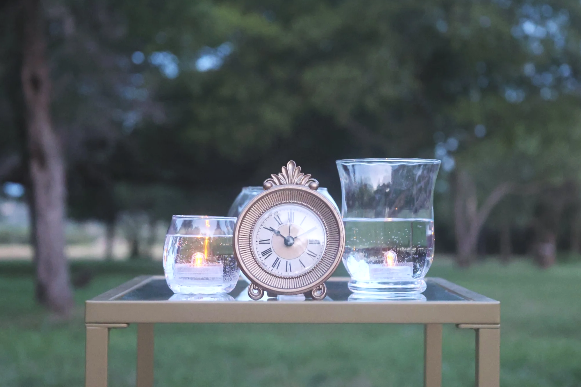 A clock and candles in glass holders on a table outdoors, with trees in the background at dusk.