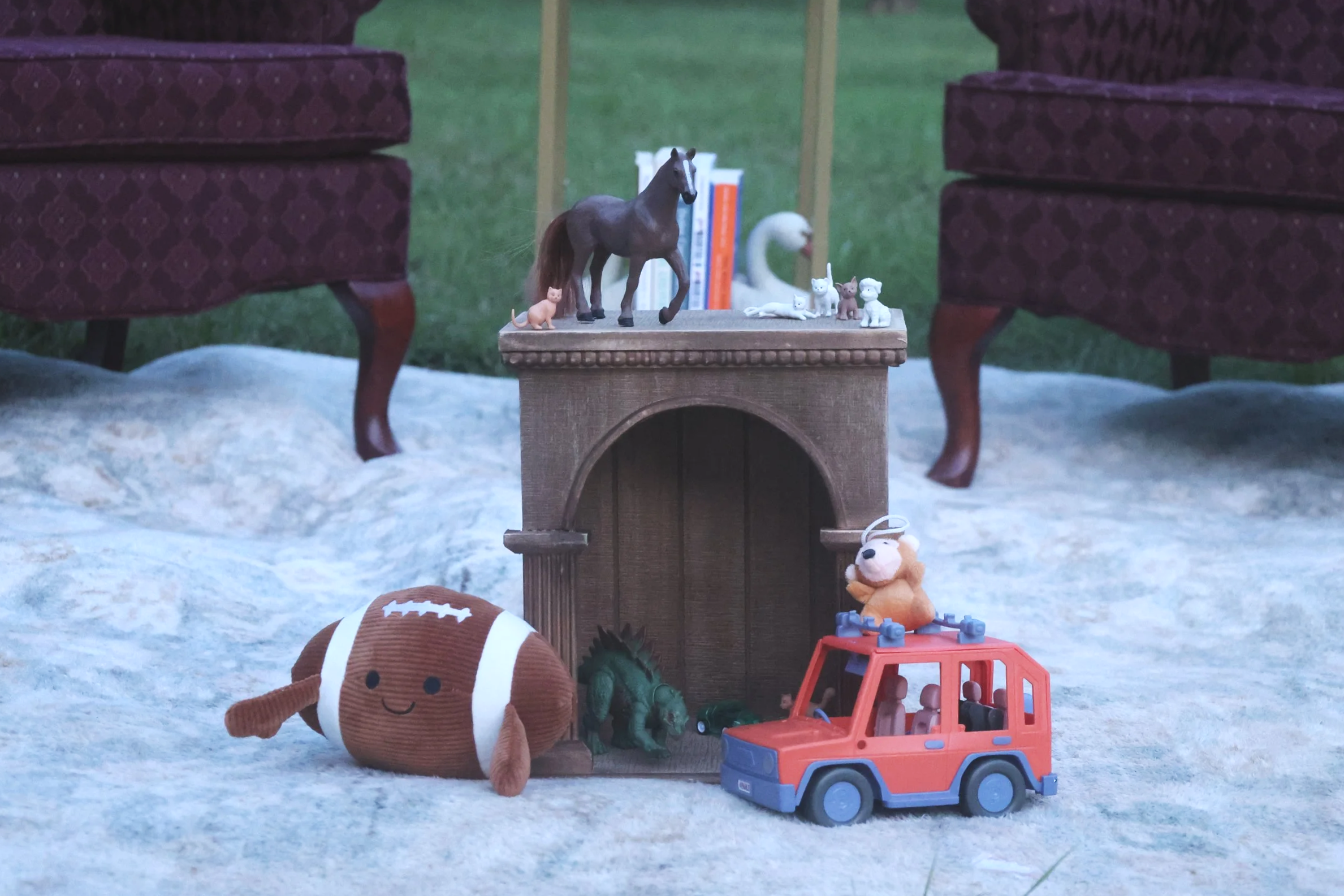Miniature toys arranged on a small table between two armchairs. The toys include a horse, several small animals, a red toy vehicle, and a plush dog. Books and a swan figurine are in the background, with a green outdoor area visible behind.