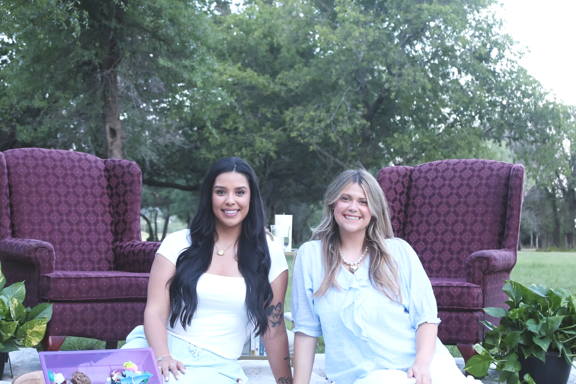 Two women sitting on the ground outdoors with large purple armchairs behind them and greenery in the background. The woman on the left has long dark hair, wearing a white top, while the woman on the right has long blonde hair, wearing a light blue shirt with a necklace. There are plants and cupcakes in front.