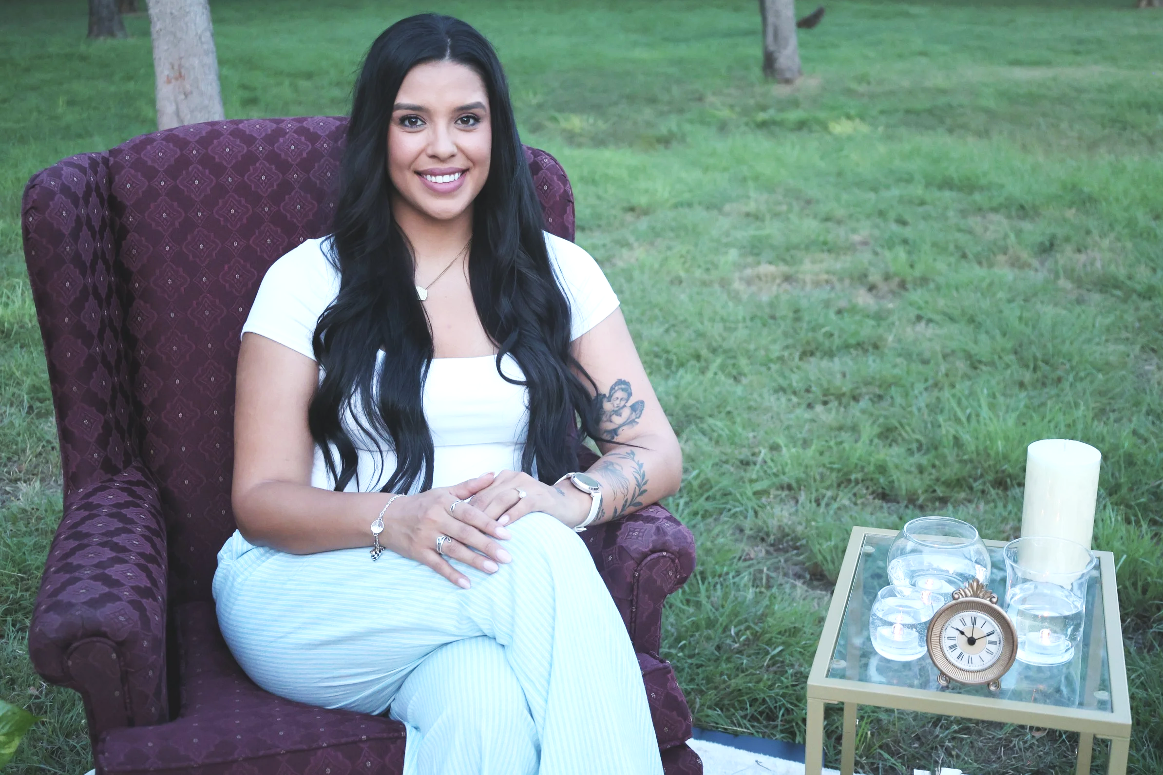 Woman with long black hair, tattoos on arms, wearing white, sitting on a purple patterned armchair outdoors, smiling, with candles, clock, and drinks on a glass table nearby.