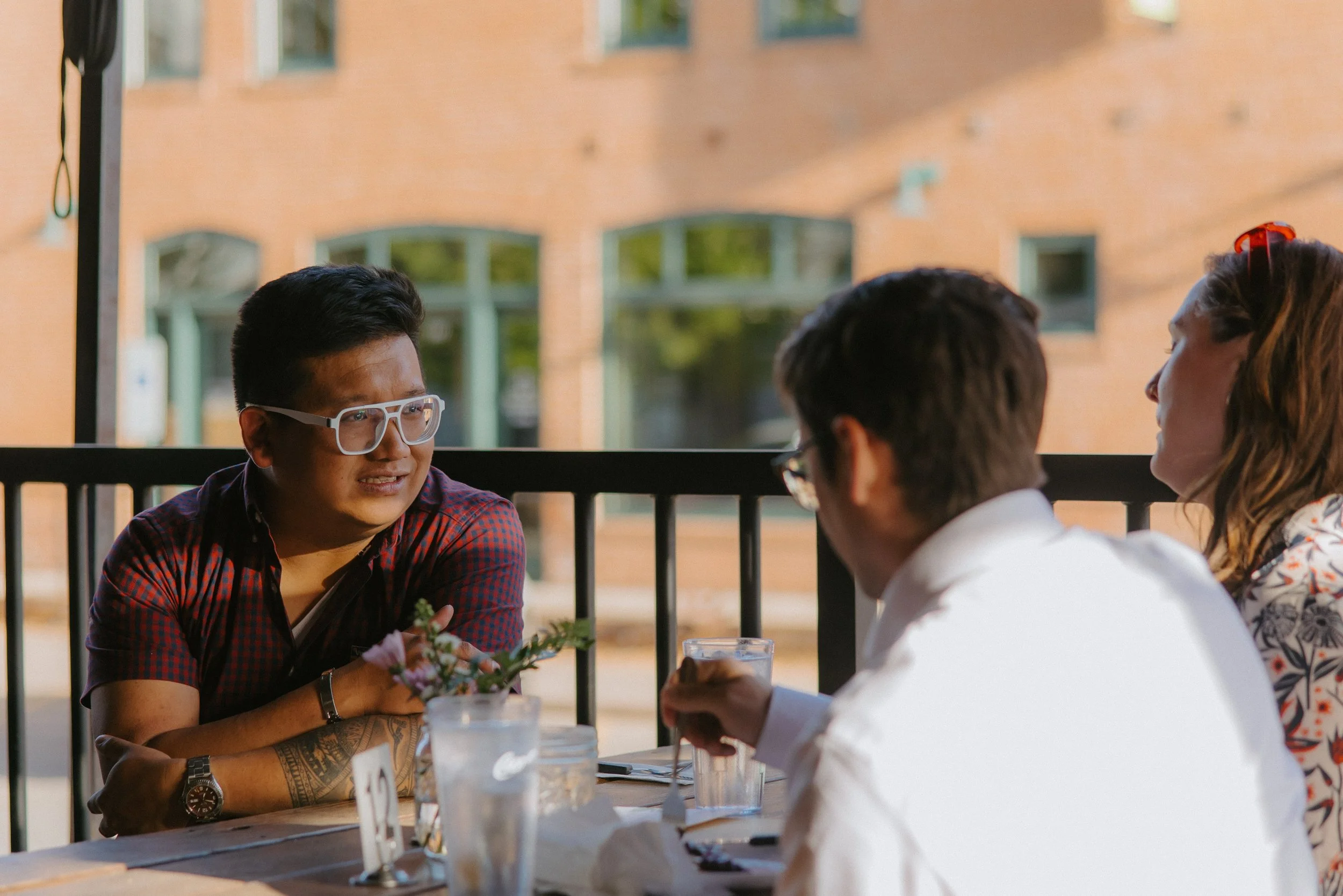 Four friends sitting at an outdoor restaurant table, engaged in conversation, with a brick building in the background.