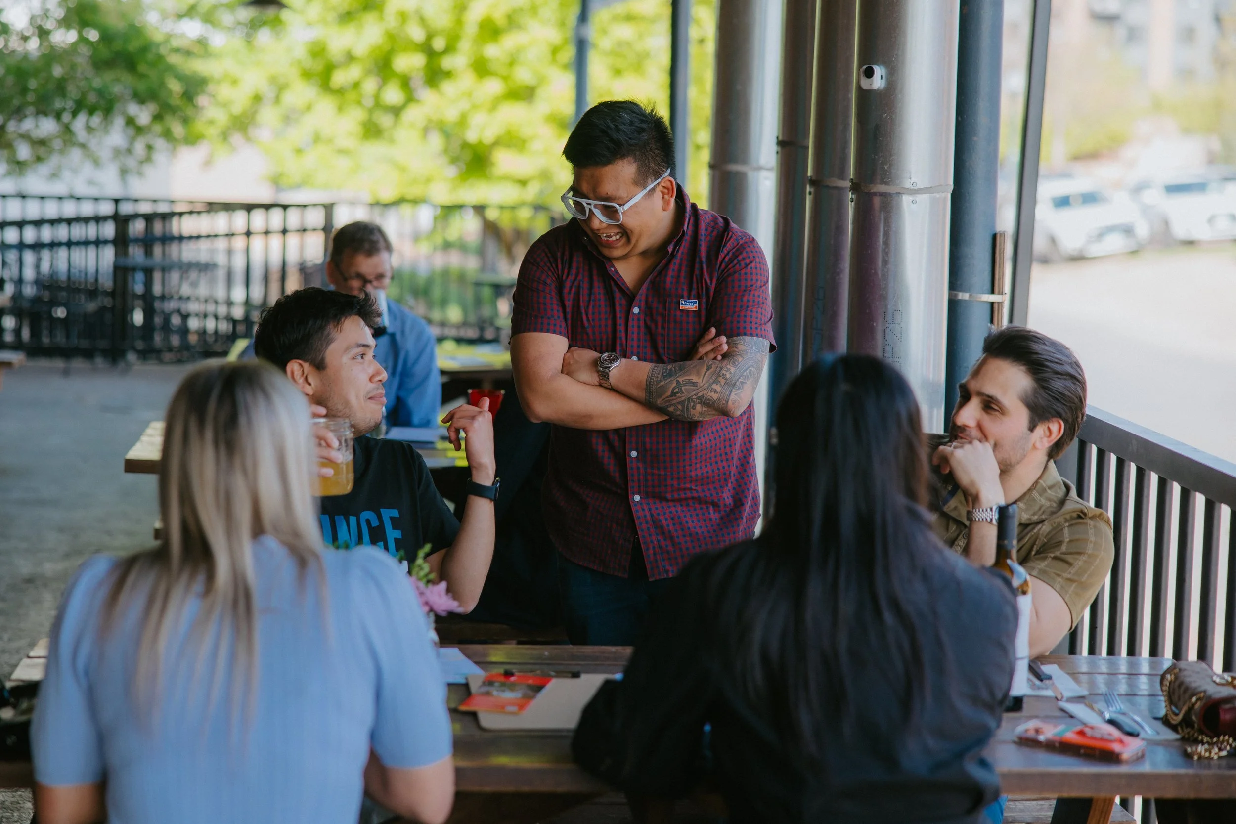 A group of people sitting at a table outside, with one man standing and smiling, engaging with others.