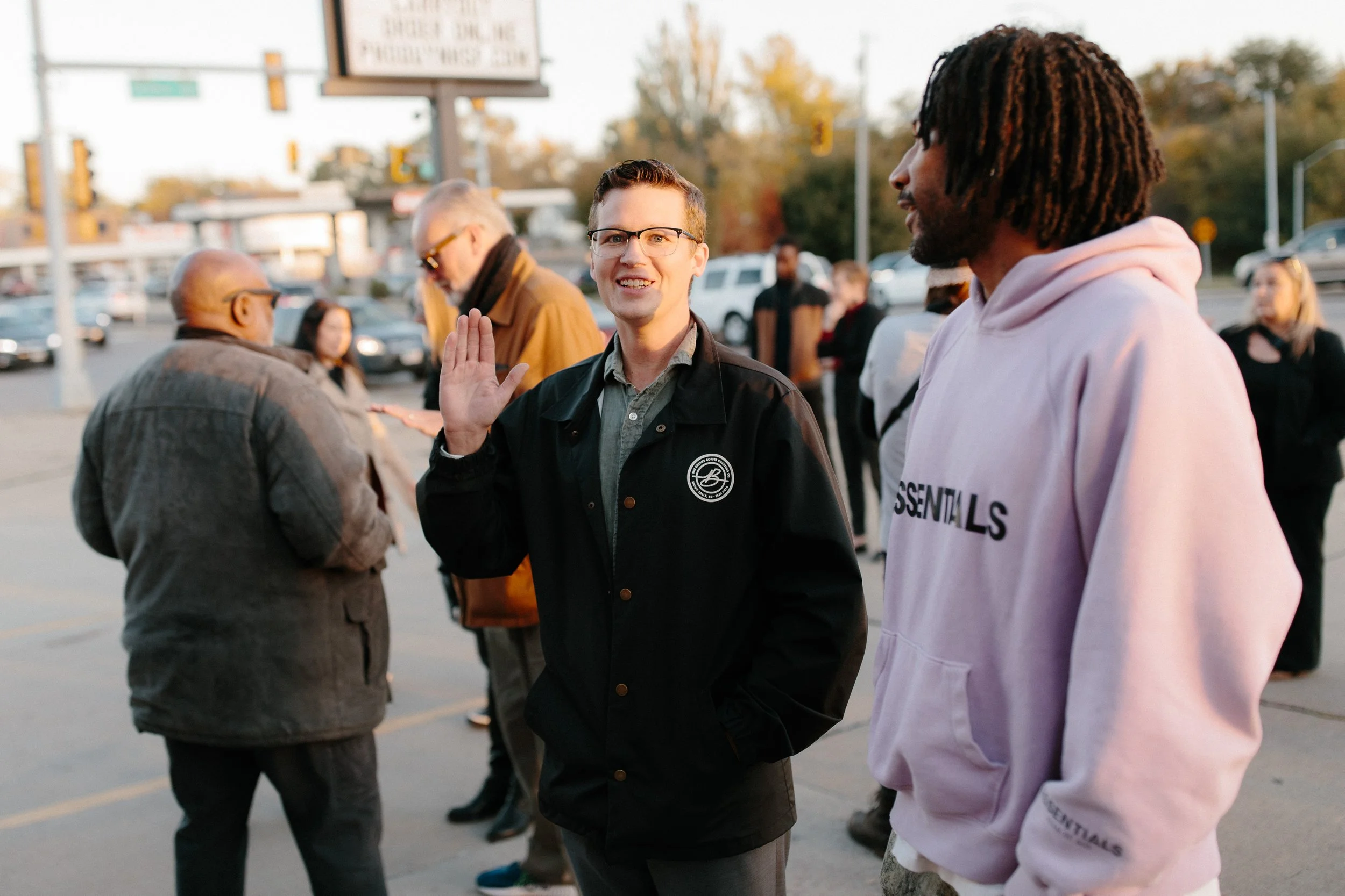 People standing outdoors in a line, with some conversing and one man in glasses waving. The background shows cars and traffic signals, suggesting a city or urban setting during the evening.