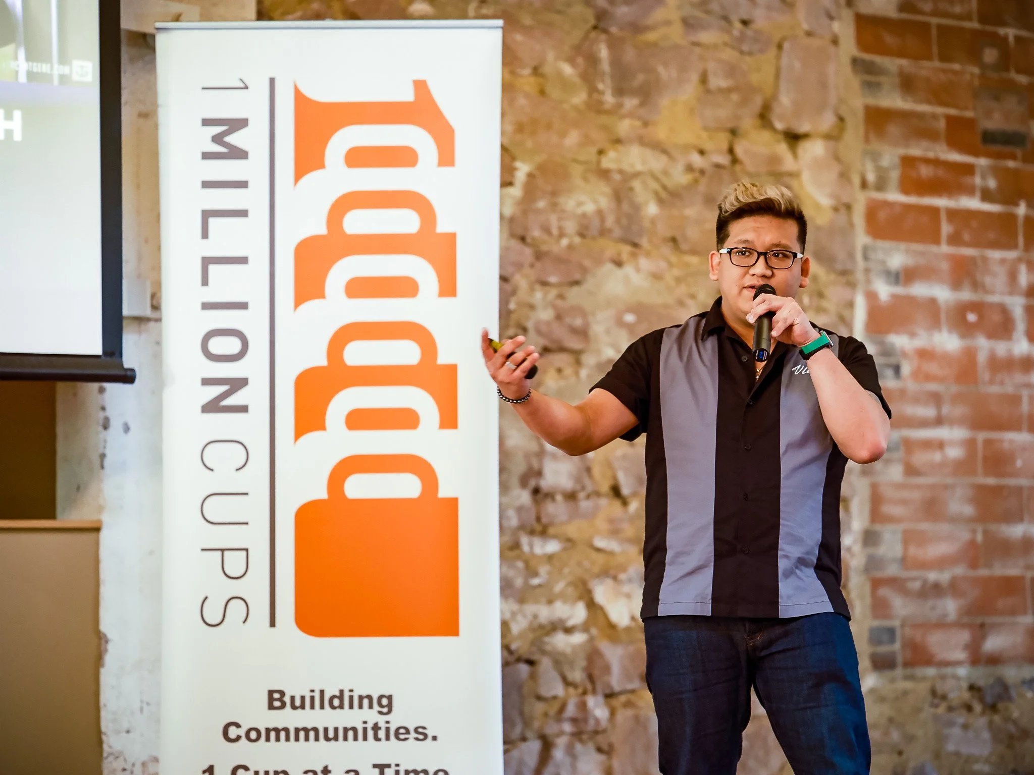 A man holding a microphone and speaking, standing next to a large vertical banner with the word 'BUILD' and other text, with a brick wall in the background.
