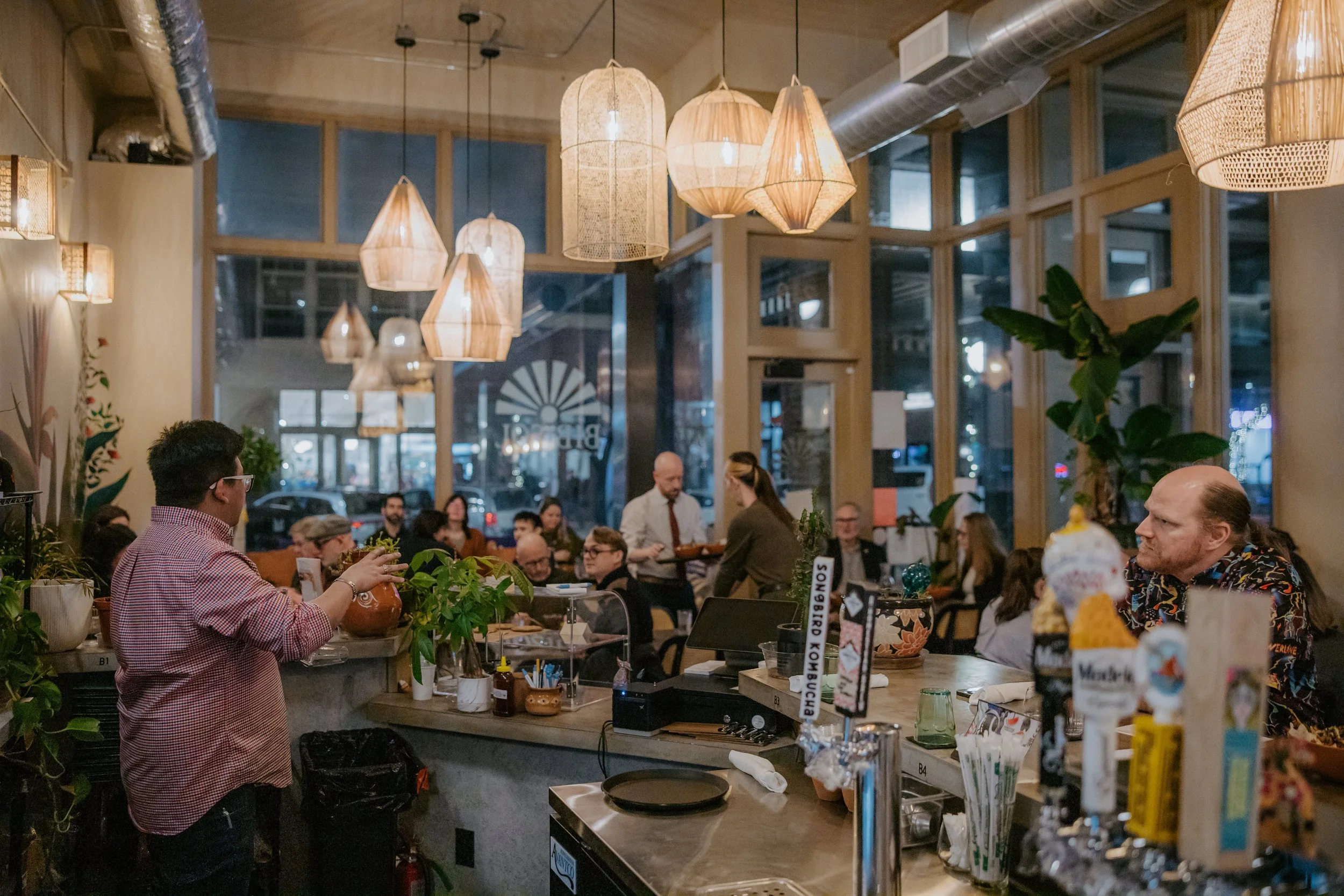 People dining and socializing inside a warmly lit restaurant with hanging pendant lights and large windows.