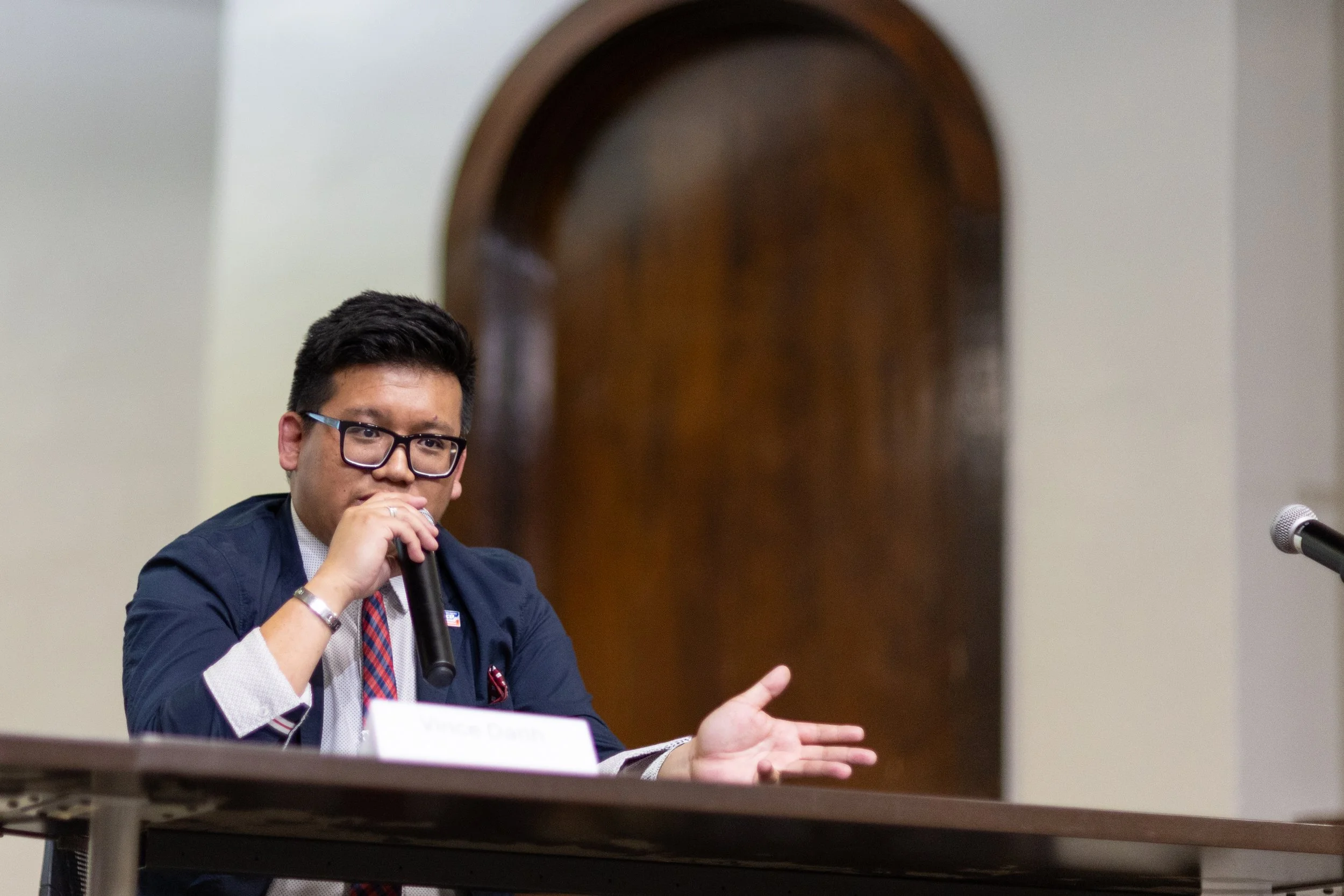 A man wearing glasses and a suit jacket speaking into a microphone during a panel discussion or meeting. There is a white nameplate in front of him that says 'Vince D...'. A microphone is positioned on the table. The background features a wooden arch and a plain wall.