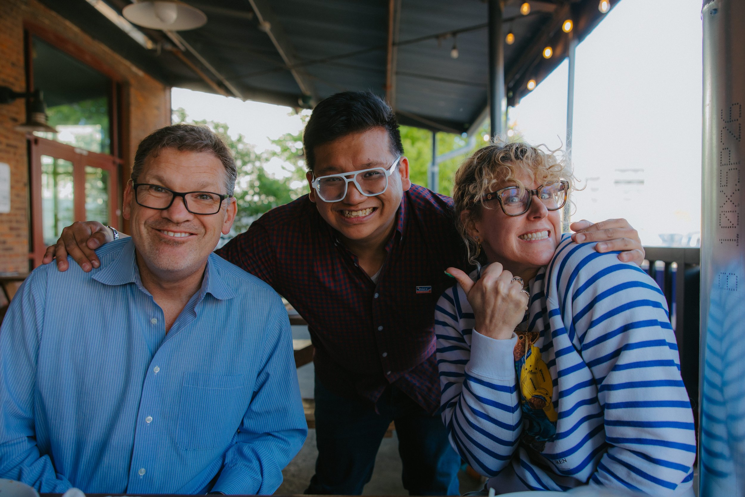 Three friends are laughing together at an outdoor cafe, with the middle person leaning close to the woman on the right. The man on the left has gray hair, glasses, and a blue shirt. The man in the middle has black hair and glasses, wearing a dark shirt. The woman on the right has curly blonde hair, glasses, and a blue and white striped shirt.