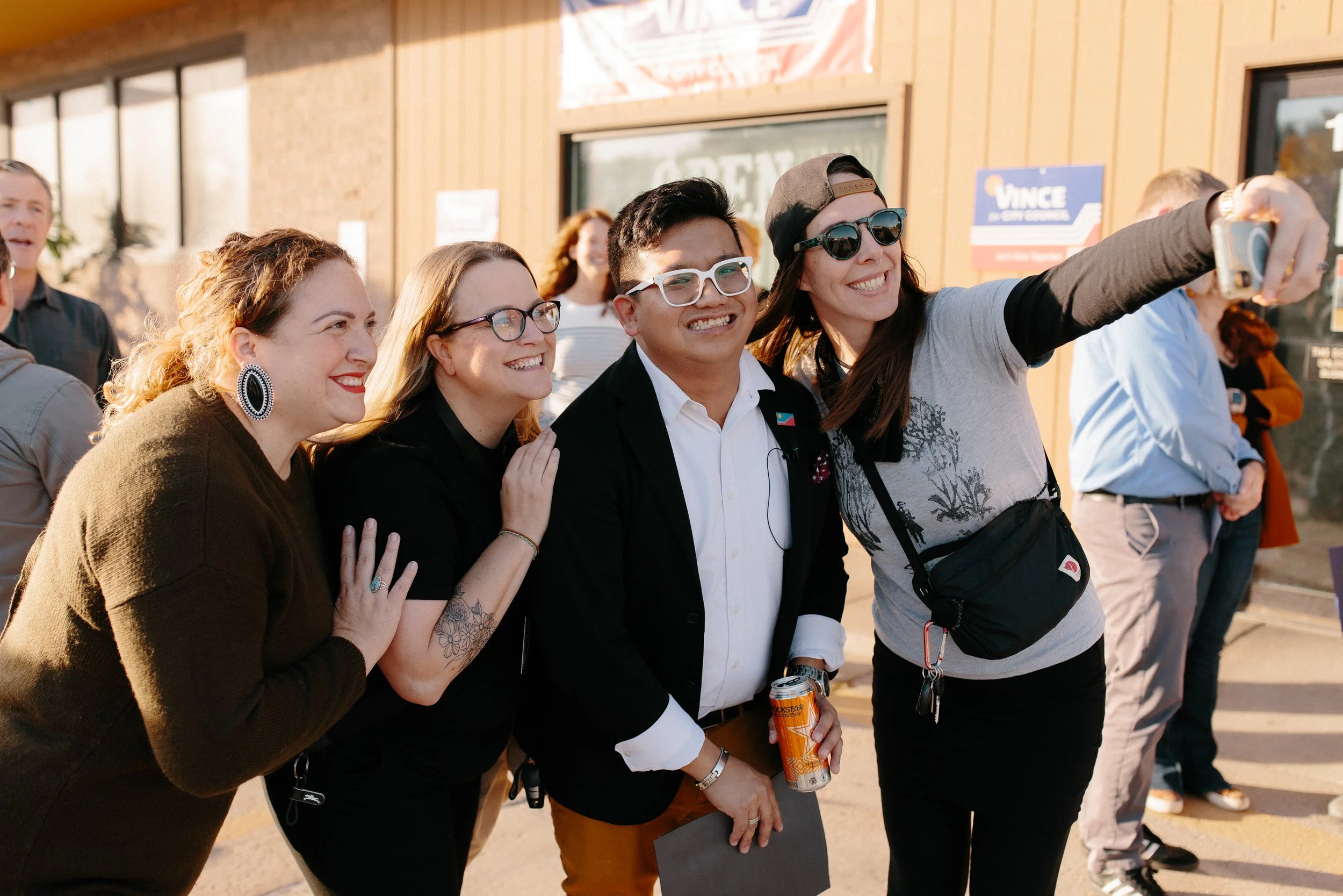 A group of five diverse people smiling and taking a selfie outdoors on a sunny day.