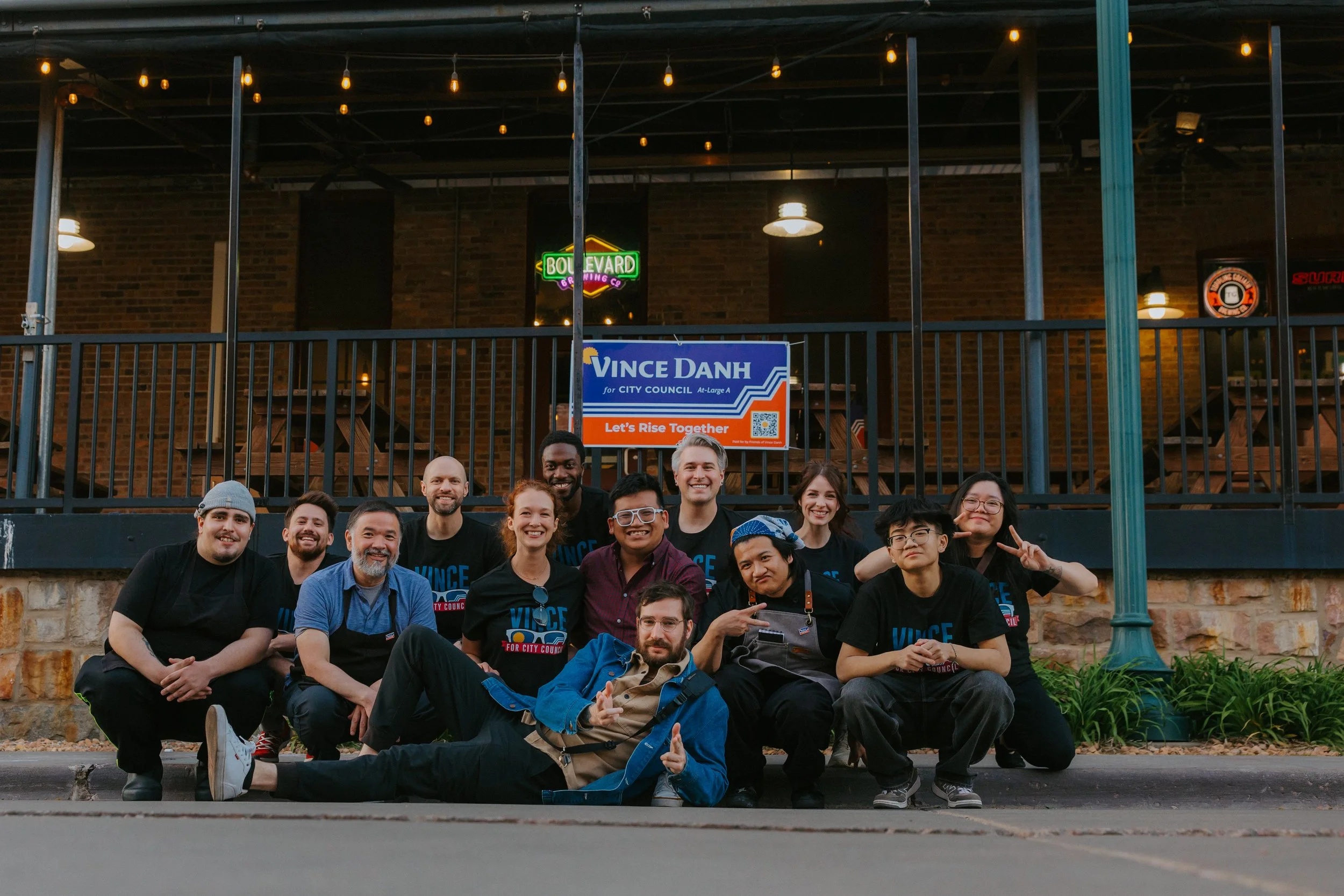 Group of people smiling and sitting on the ground in front of a building with neon signs and political campaign signs during evening.