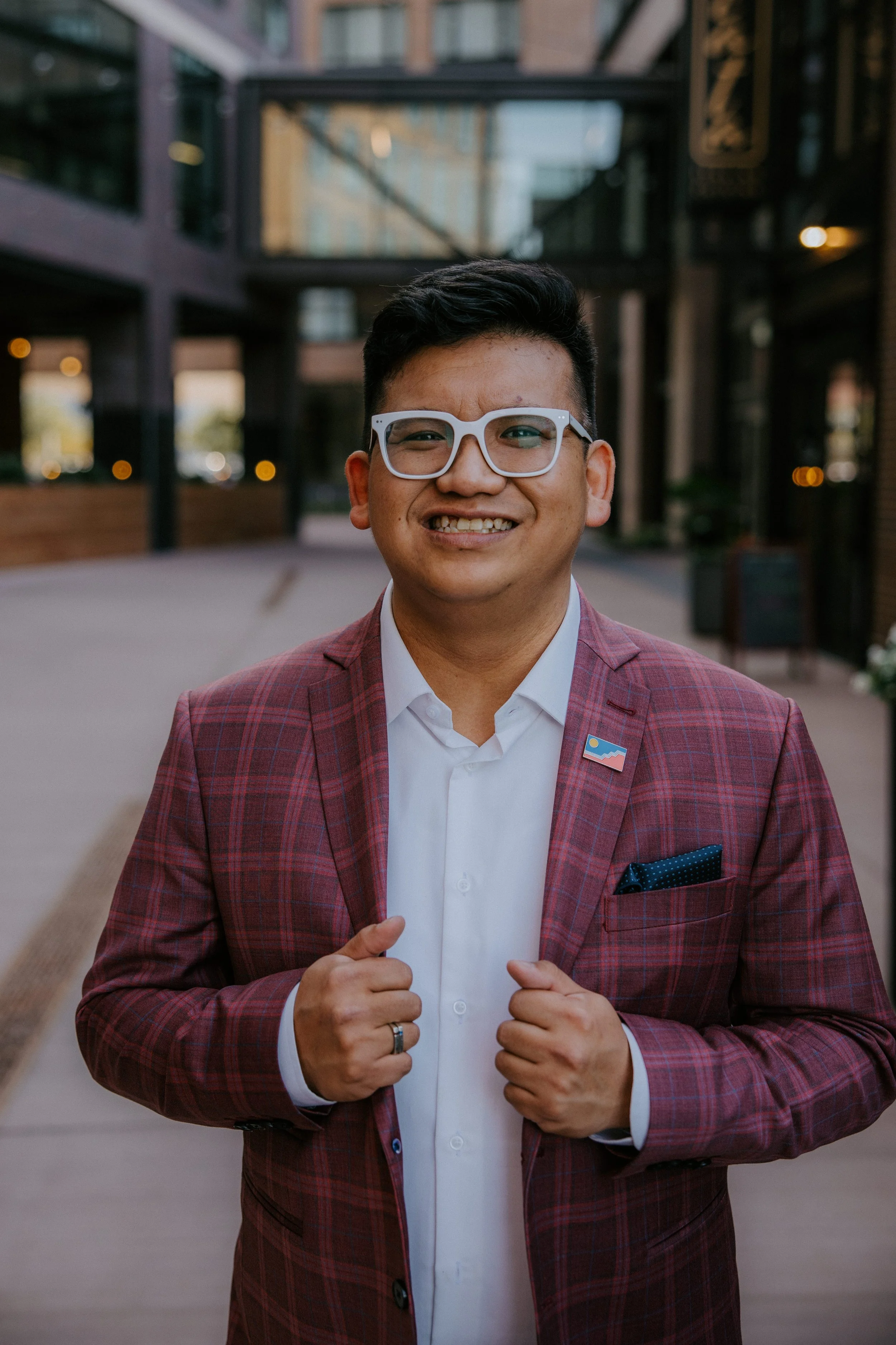 A man in a red plaid suit and white shirt, wearing glasses, smiling and holding the lapels of his suit jacket. He is outdoors in an urban setting with modern buildings in the background.