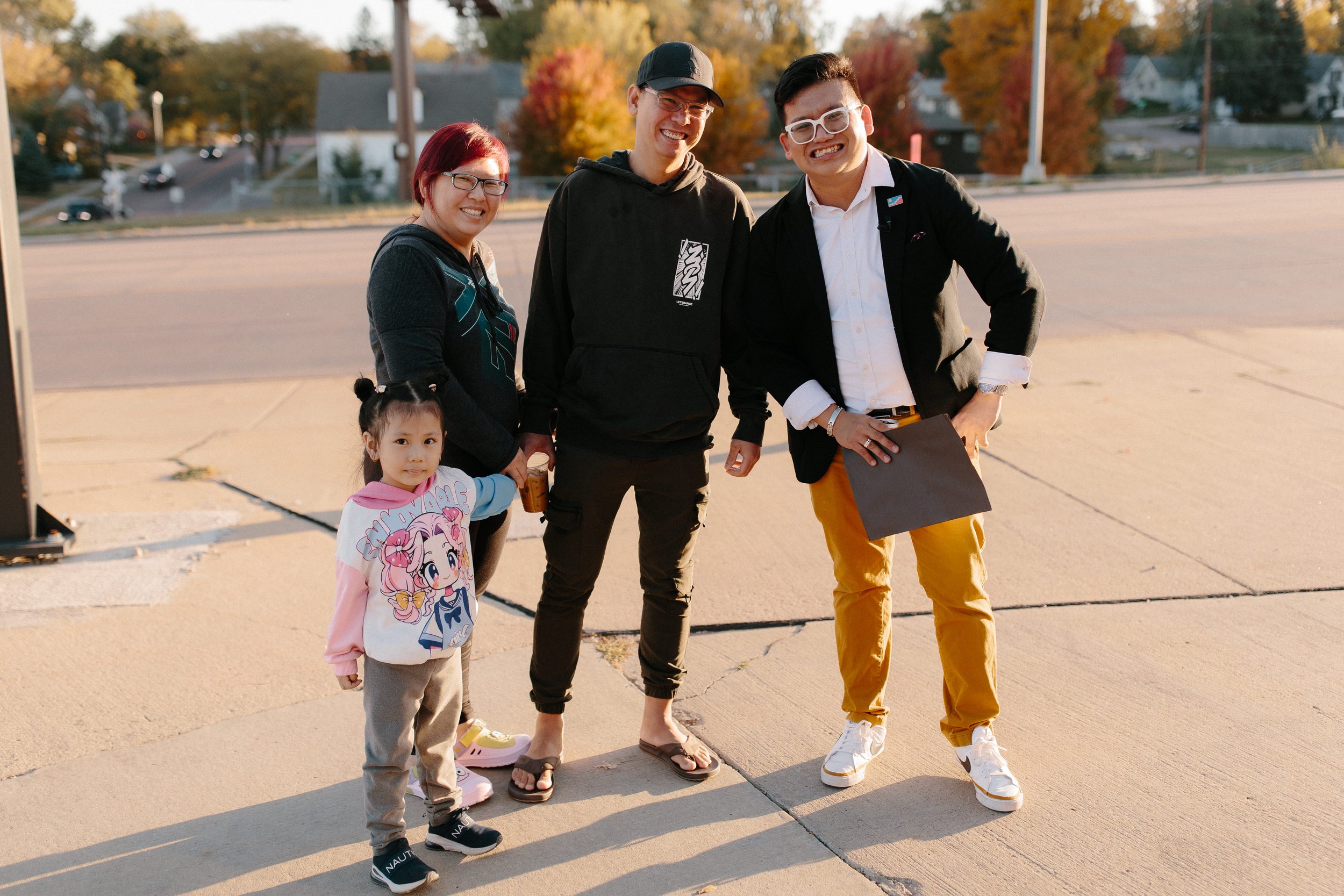 Four people, including a young girl, posing happily outdoors during autumn, with trees showing fall colors and a suburban neighborhood in the background.