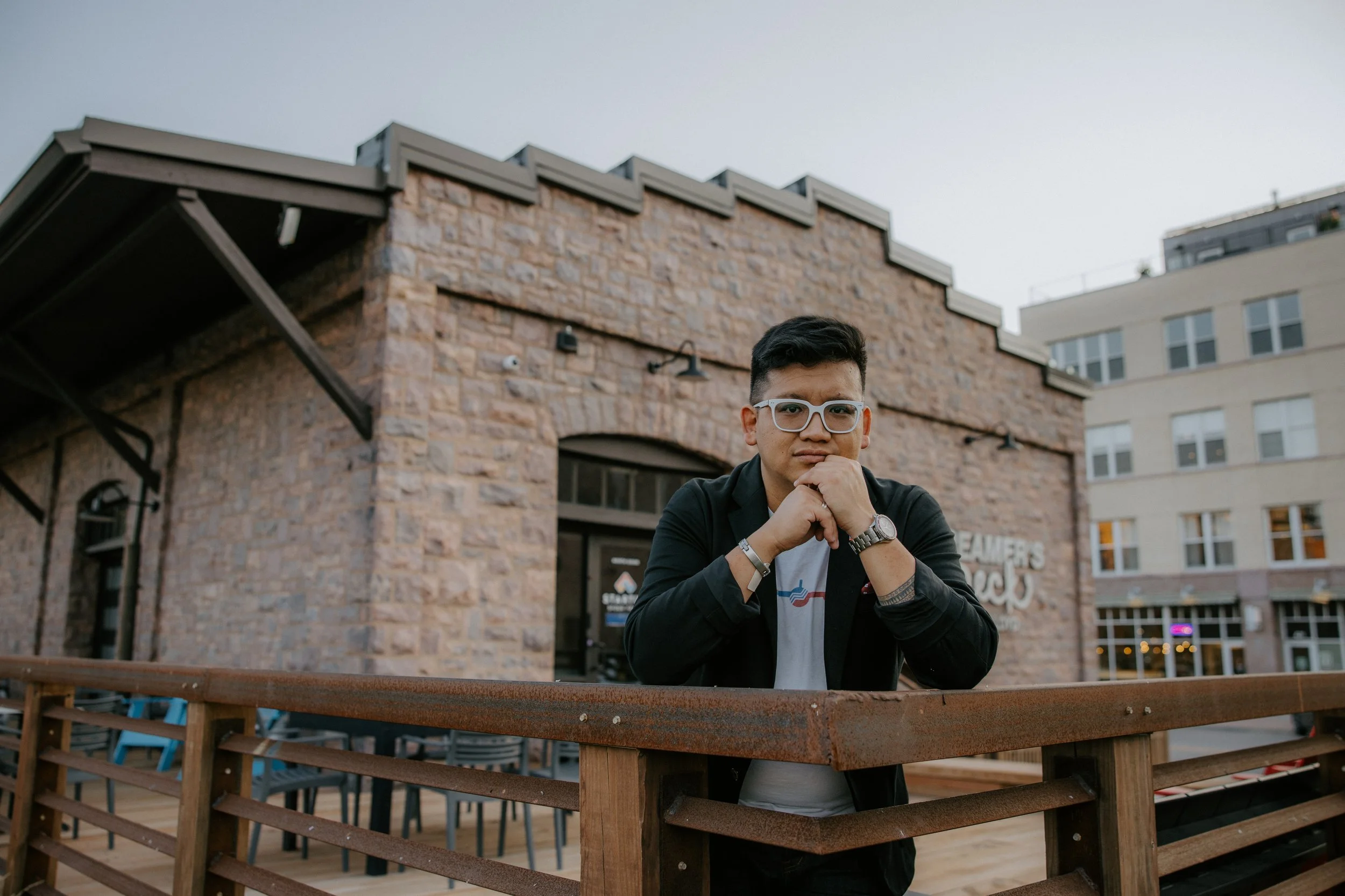 A young man with glasses and dark hair sits outdoors with his chin resting on his hand, in front of a brick building with string lights, on a cloudy day.