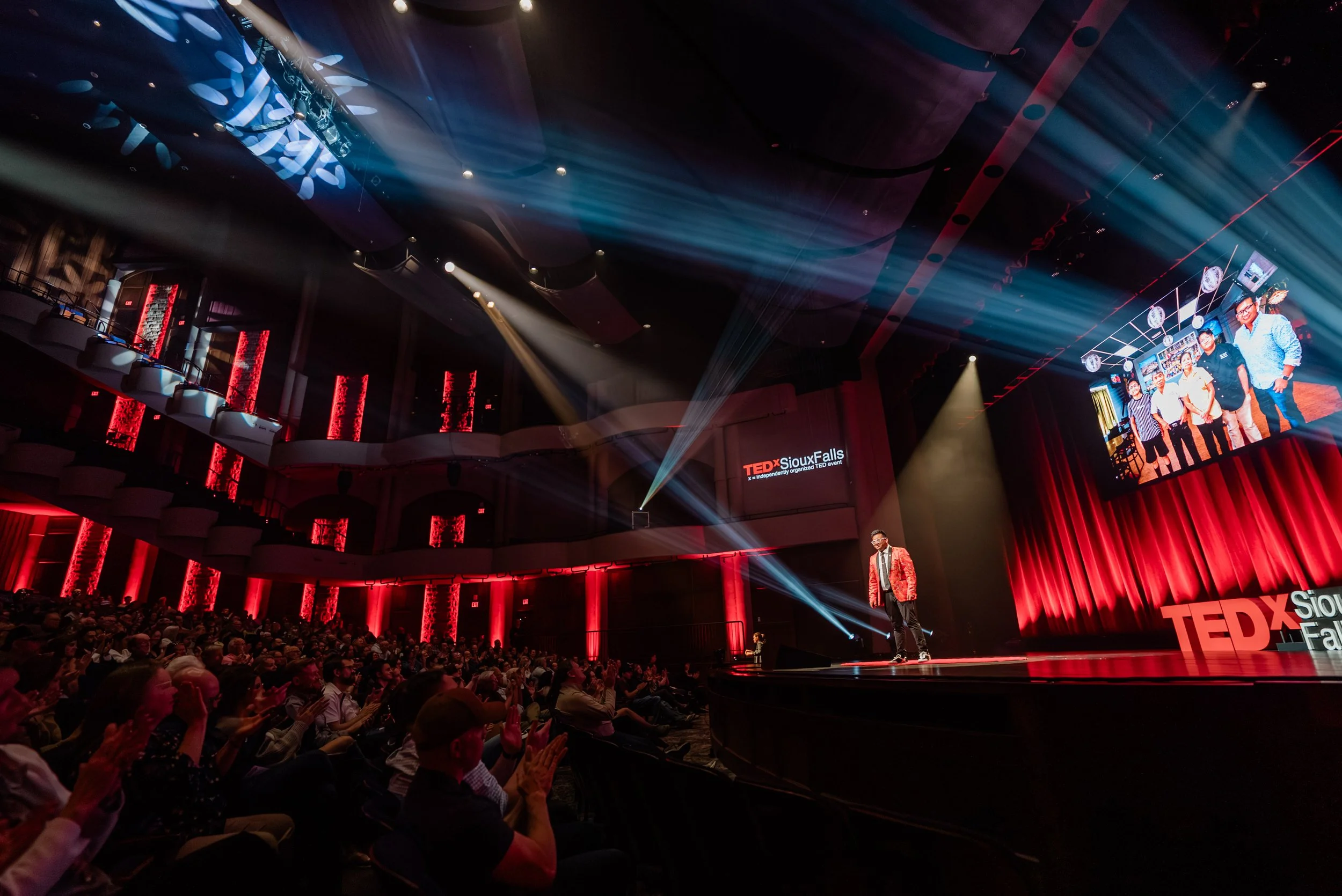 An auditorium filled with people attending a TEDx event at Sioux Falls. The stage is lit with blue and red lights, with a person speaking on stage and a large screen displaying a group photo of five people.
