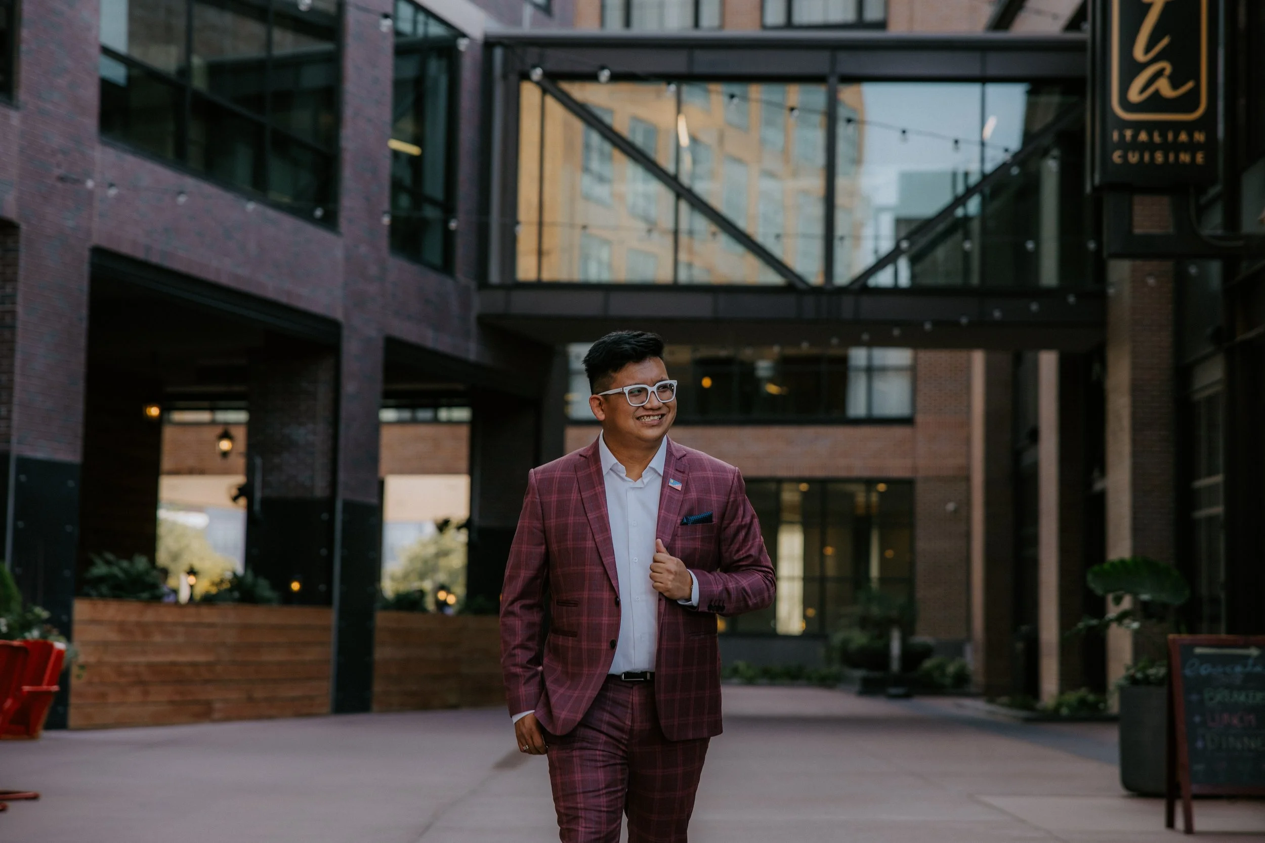 A man with glasses and a short haircut, wearing a pink plaid suit and white shirt, walking outside in an urban area with brick and glass buildings, smiling.