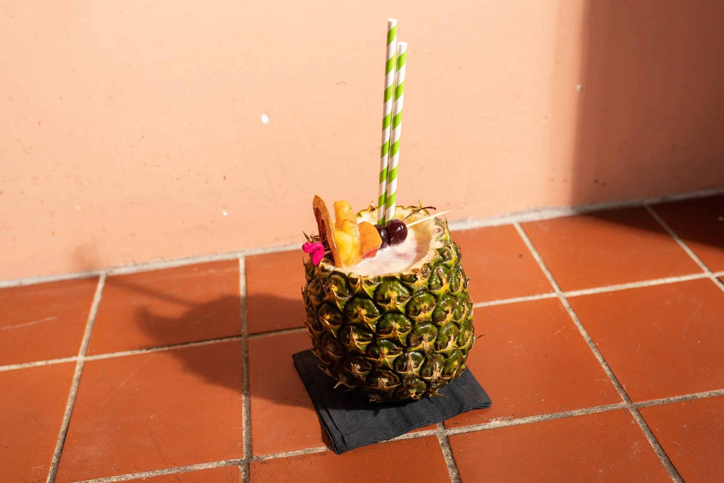 A pineapple used as a cup with a tropical beverage inside, topped with fruit and a straw, placed on a black napkin on a tiled floor.