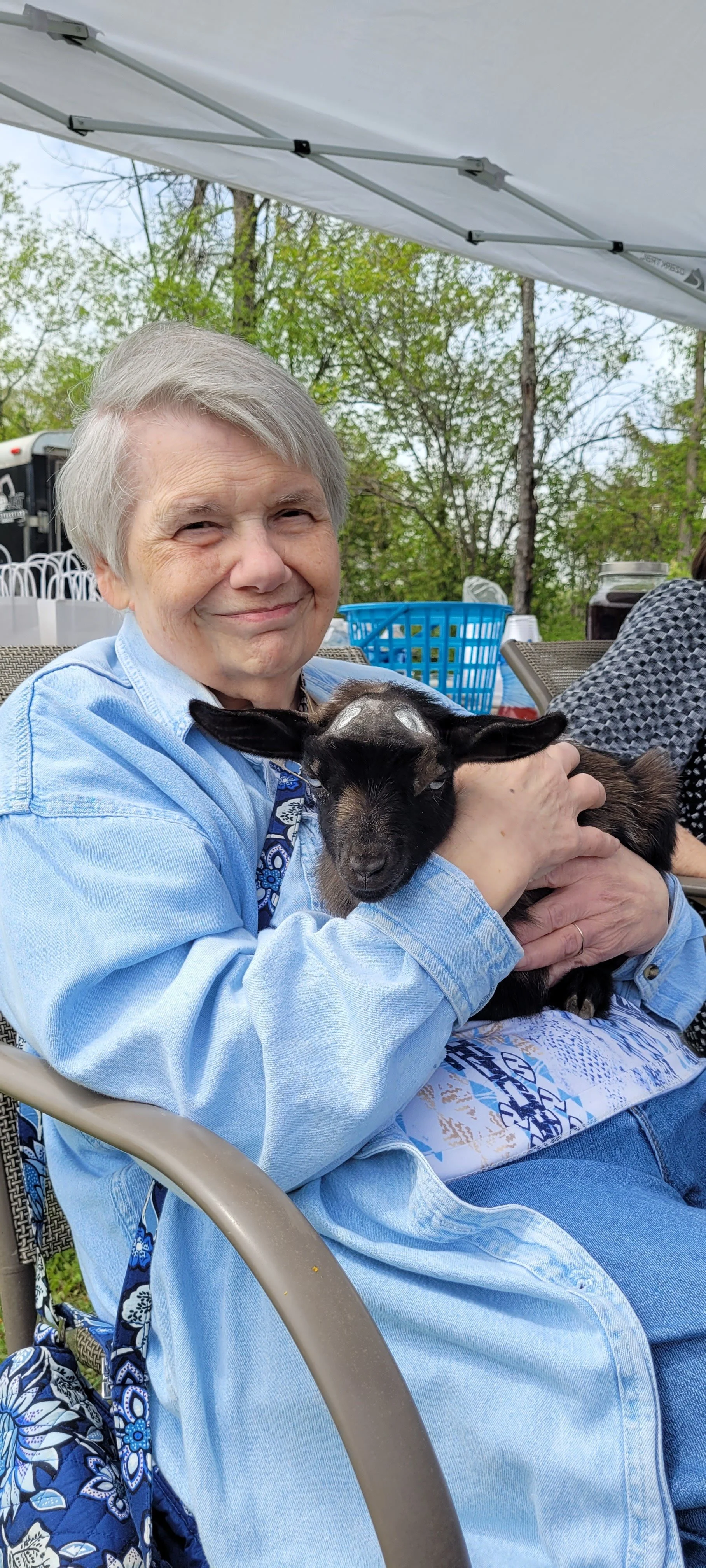 An elderly woman with short gray hair smiling and holding a small black and brown goat in her arms outdoors under a white canopy, with trees and other seated people in the background.
