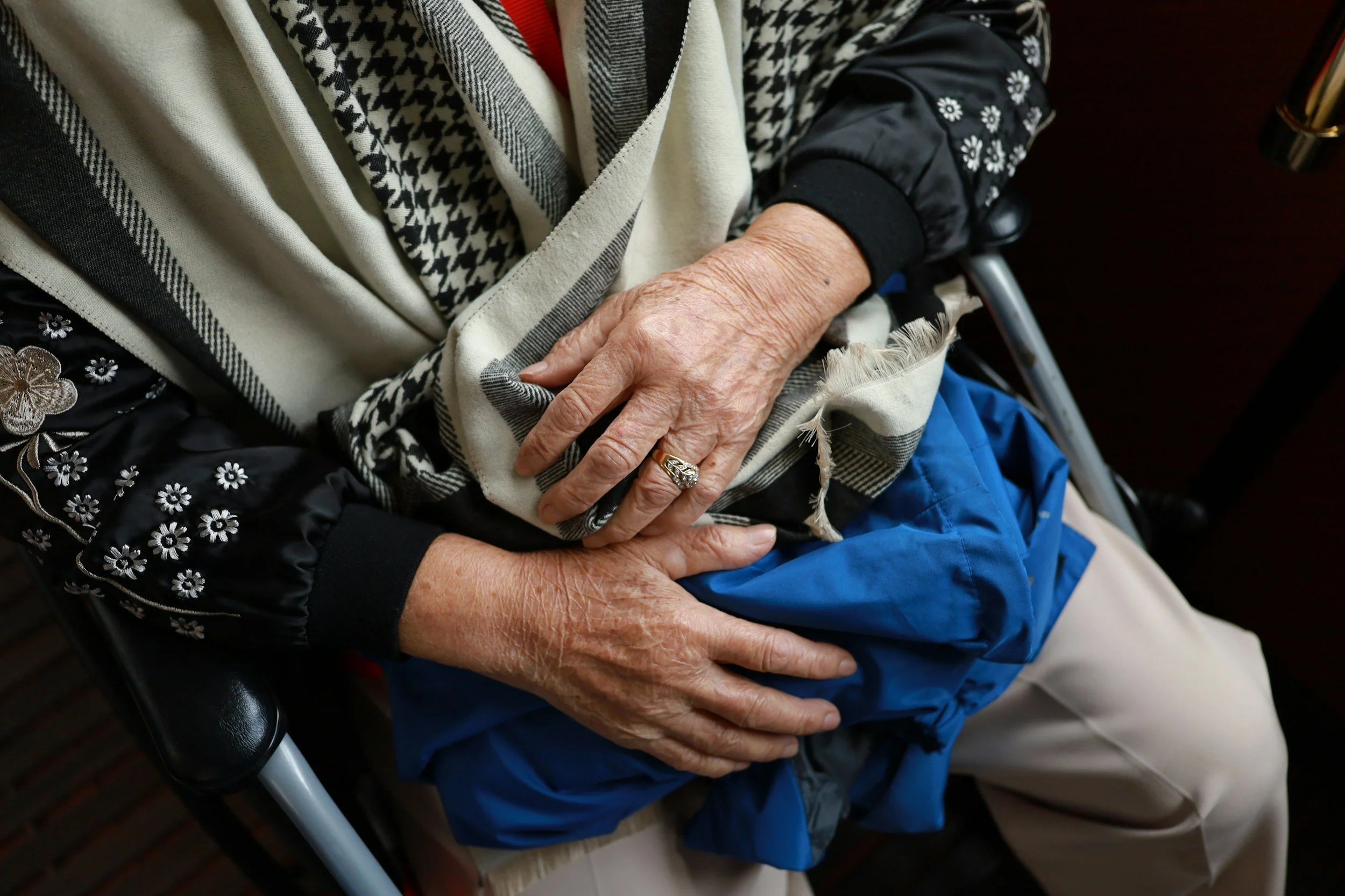 Close-up of an elderly person's hands resting on their lap, wearing a silver ring and a plaid scarf, seated in a wheelchair.