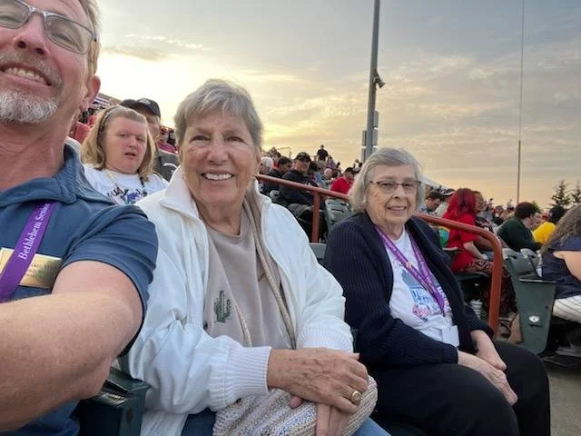 People sitting in stadium bleachers, watching an outdoor event during sunset. Focus on three smiling individuals, with a man on the left partly visible and two elderly women in the center and right.