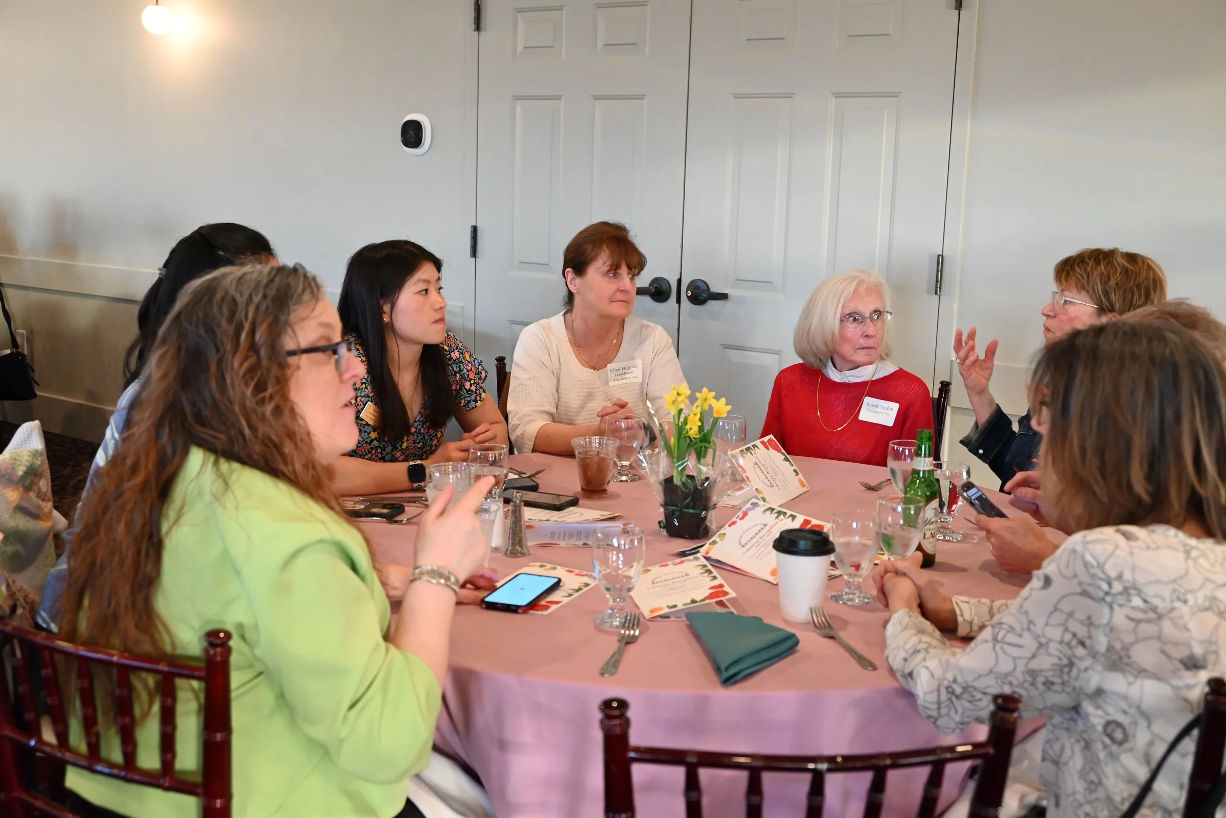 Women sitting around a banquet table with pink tablecloth, various glasses, menus, and a vase with yellow flowers, engaged in conversation at a social gathering or dinner event.