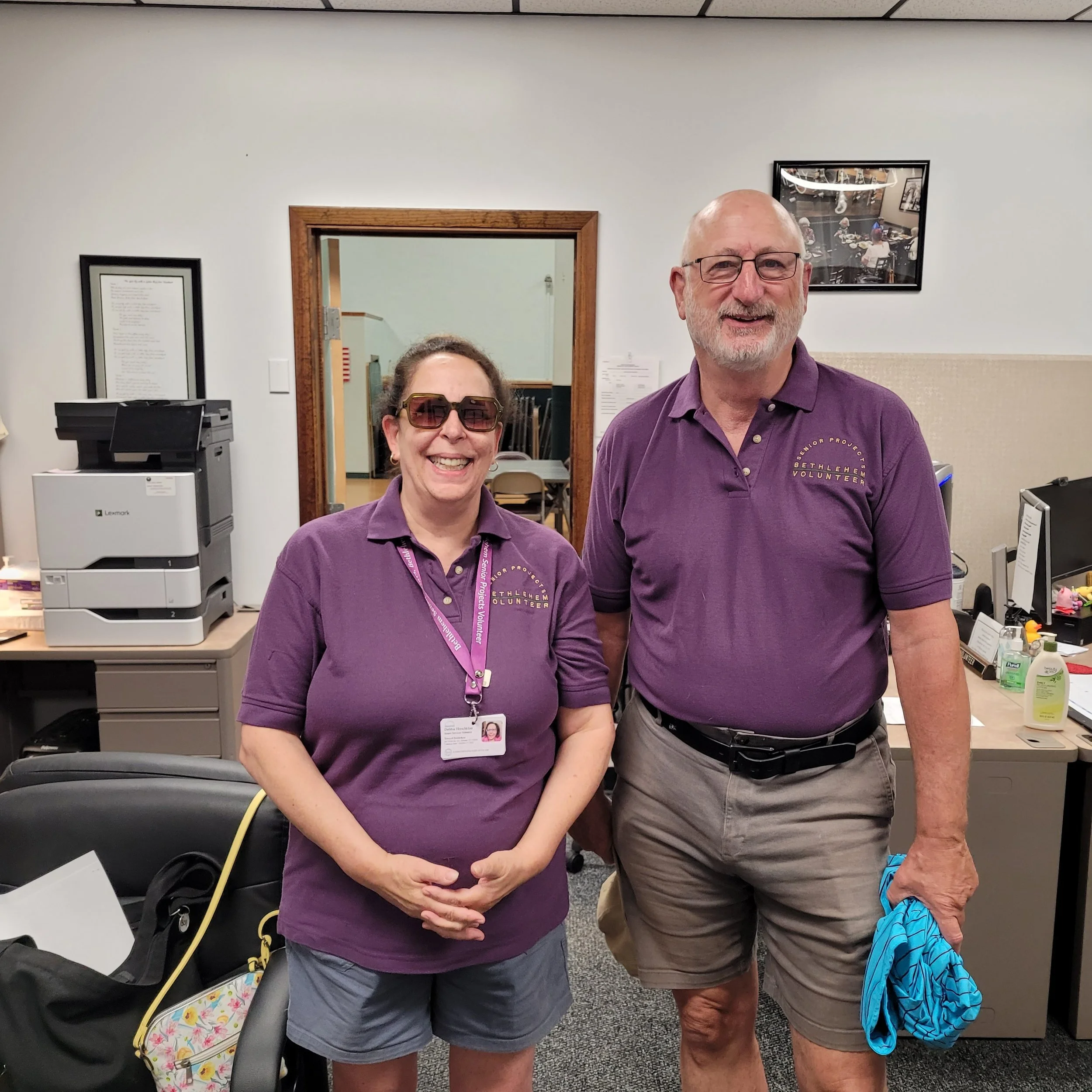 Two smiling individuals, a woman and a man, standing in an office or reception area. They are wearing matching purple shirts with volunteer or project logos. The woman has sunglasses and a name badge, and the man has glasses. Behind them are office s