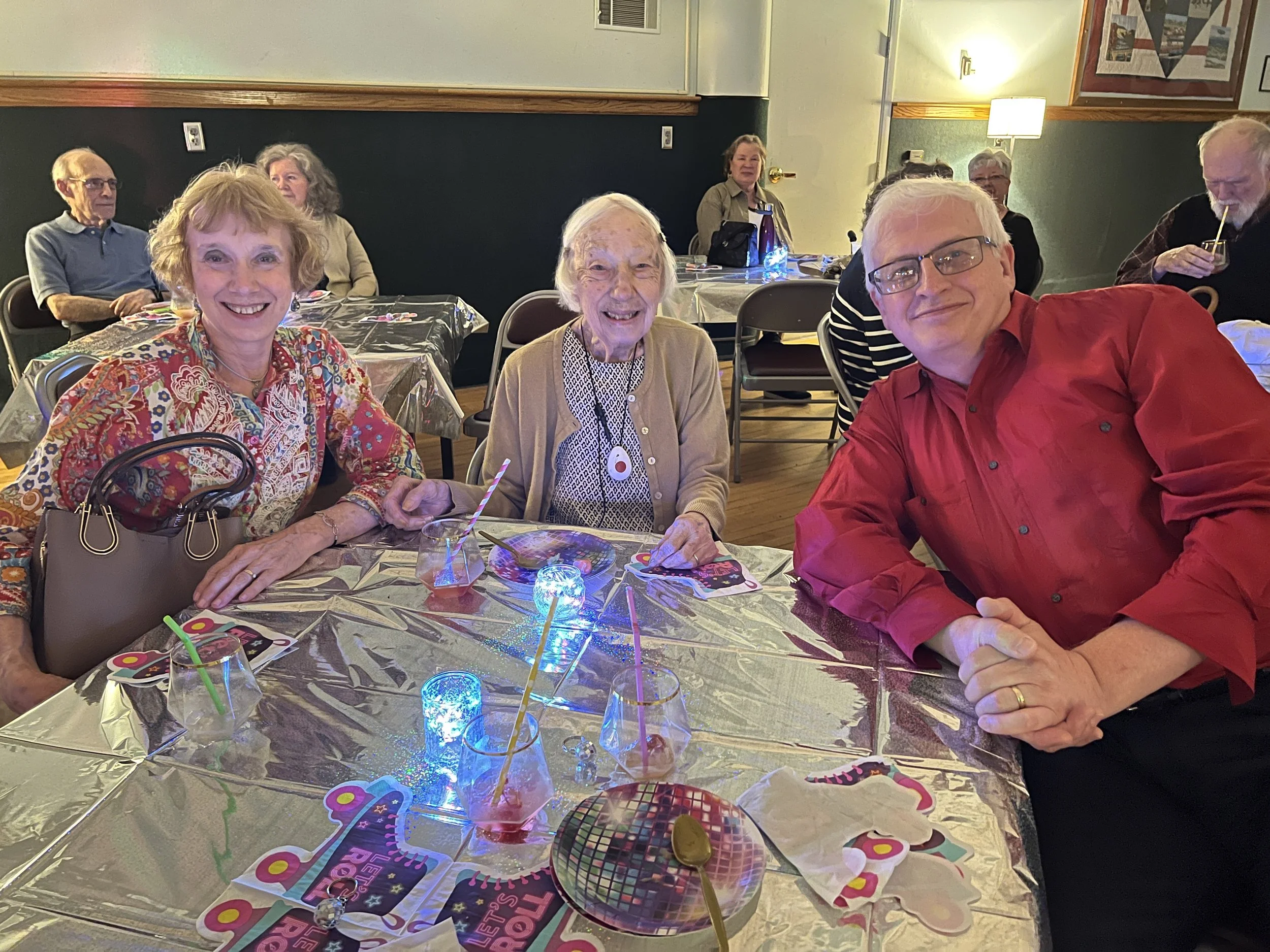 A group of older adults sitting at a table during a celebration, with colorful party decorations, confetti, and birthday-themed items, smiling and enjoying the event.