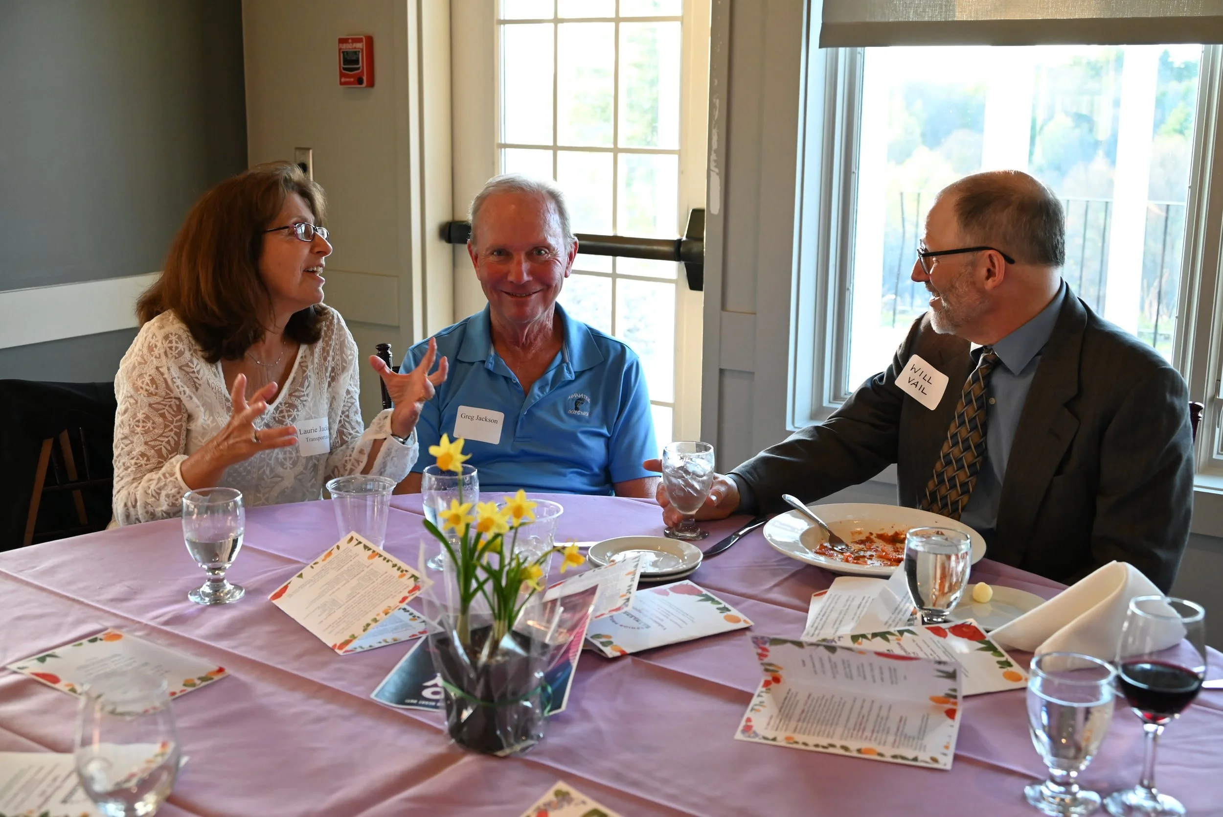 Three adults sitting at a table in a bright room, engaged in conversation. The table has a pink tablecloth, menus, and a small flower arrangement with yellow flowers. One man is smiling and wearing a blue polo shirt with a name tag that says Greg Jac