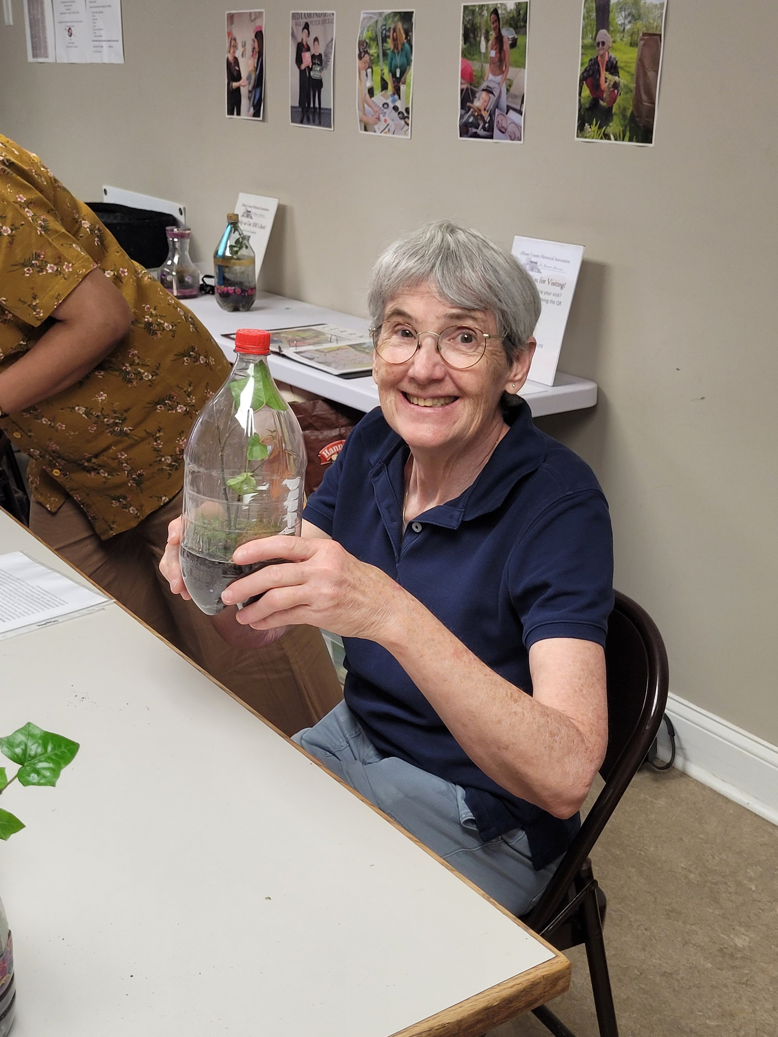 An elderly woman with short gray hair and glasses smiling and holding a clear plastic water bottle filled with water and green leaves. She is seated at a table in a room with a beige wall, decorated with photographs of people, and informational signs