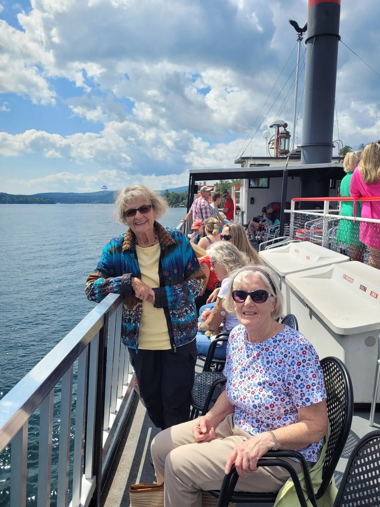 A group of senior women enjoying a boat ride on a lake, with some sitting and others standing, under a partly cloudy sky.