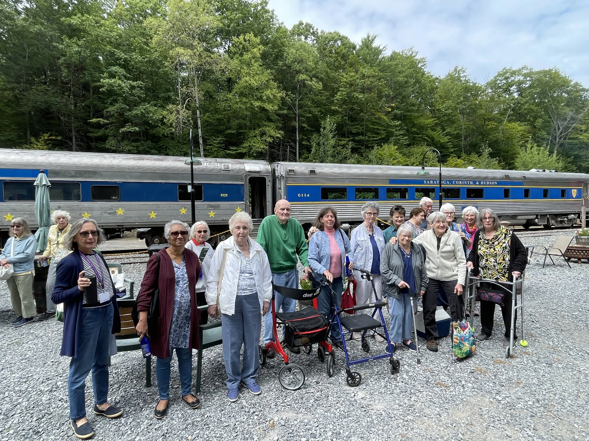 A group of elderly people standing outdoors near a blue train car, with some carrying walkers and bags, in front of a forested area with green trees and cloudy sky.