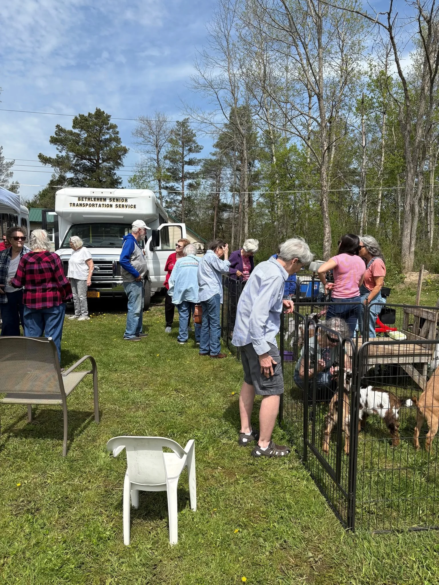 A group of people gathered outdoors near a fenced area with dogs, behind a white van labeled 'Bethlehem Senior Transportation Service', in a park-like setting with trees and a blue sky.