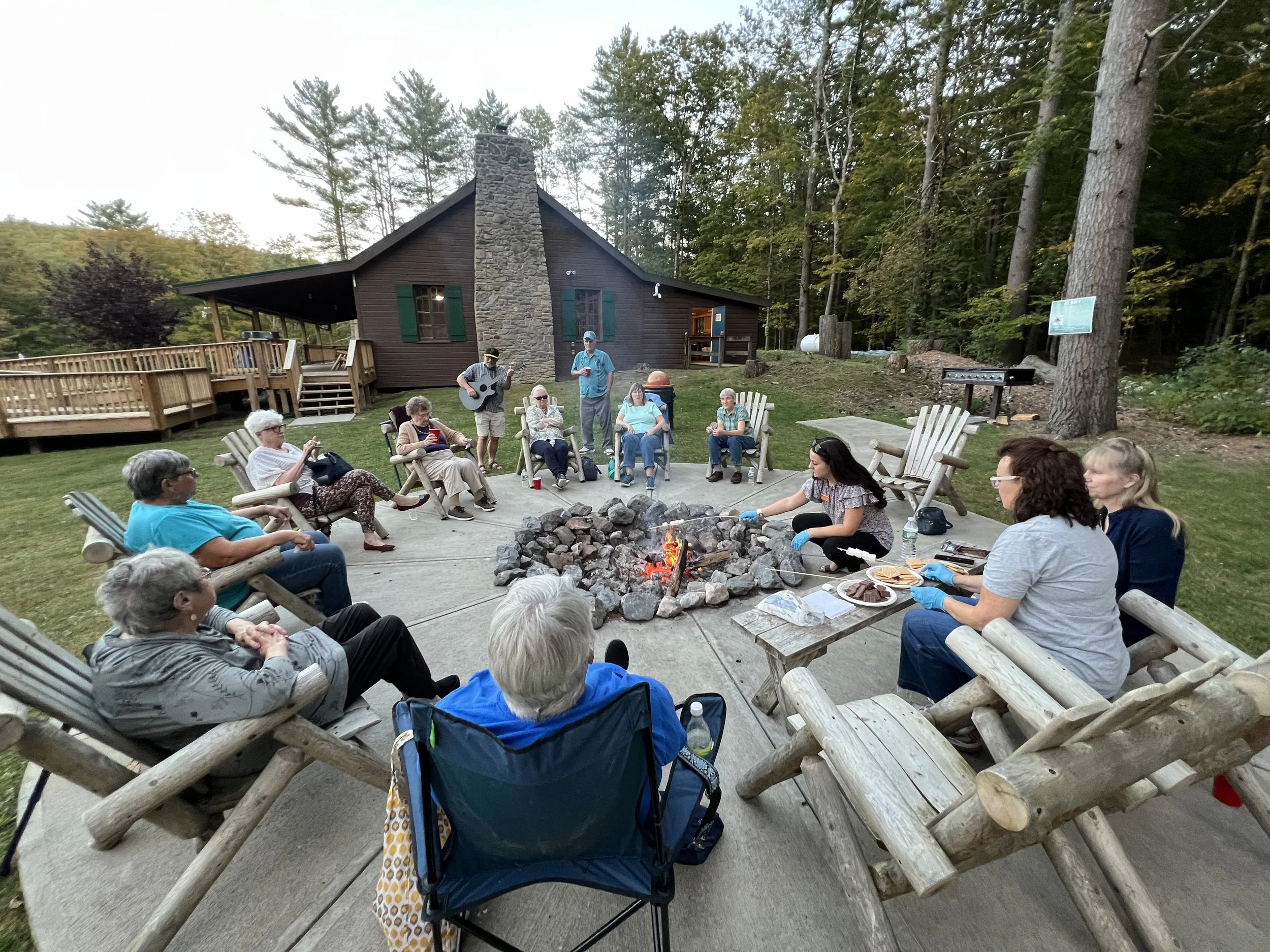 Group of people sitting around a firepit in front of a cabin with a stone chimney, surrounded by tall trees, some holding drinks and a guitarist performing, with a grill on the side.