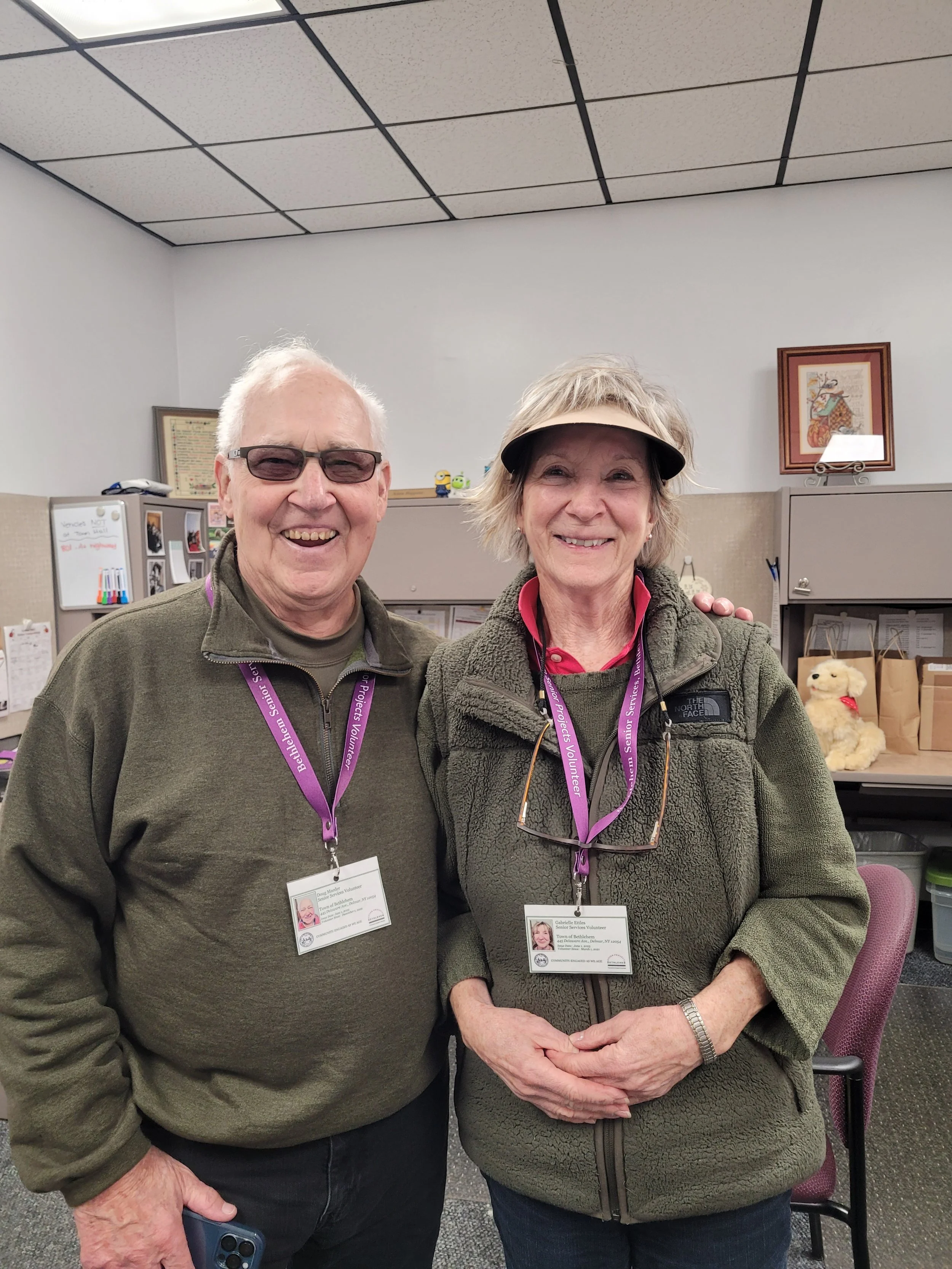 A man and a woman smiling and standing close together in an office environment, both wearing volunteer identification badges and purple lanyards. The man is wearing sunglasses and a green jacket, holding a phone, while the woman is wearing a visor and a green fleece jacket.