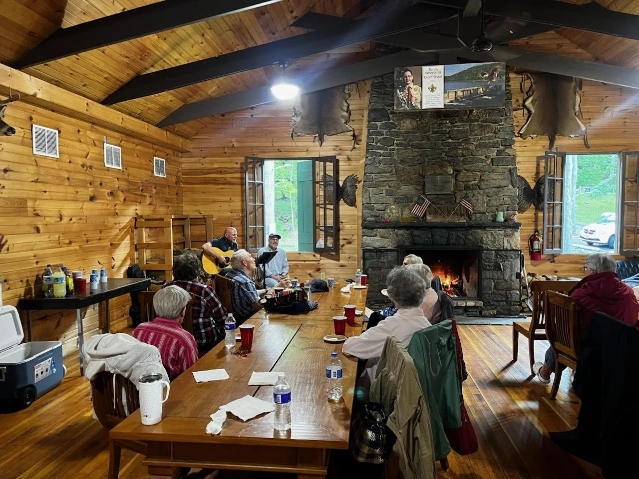 A group of elderly people gathered in a rustic wood-paneled room with a large stone fireplace. Some are sitting at tables, drinking and eating, while one person is performing with a guitar near the fireplace. The room has open windows, cowboy hats on